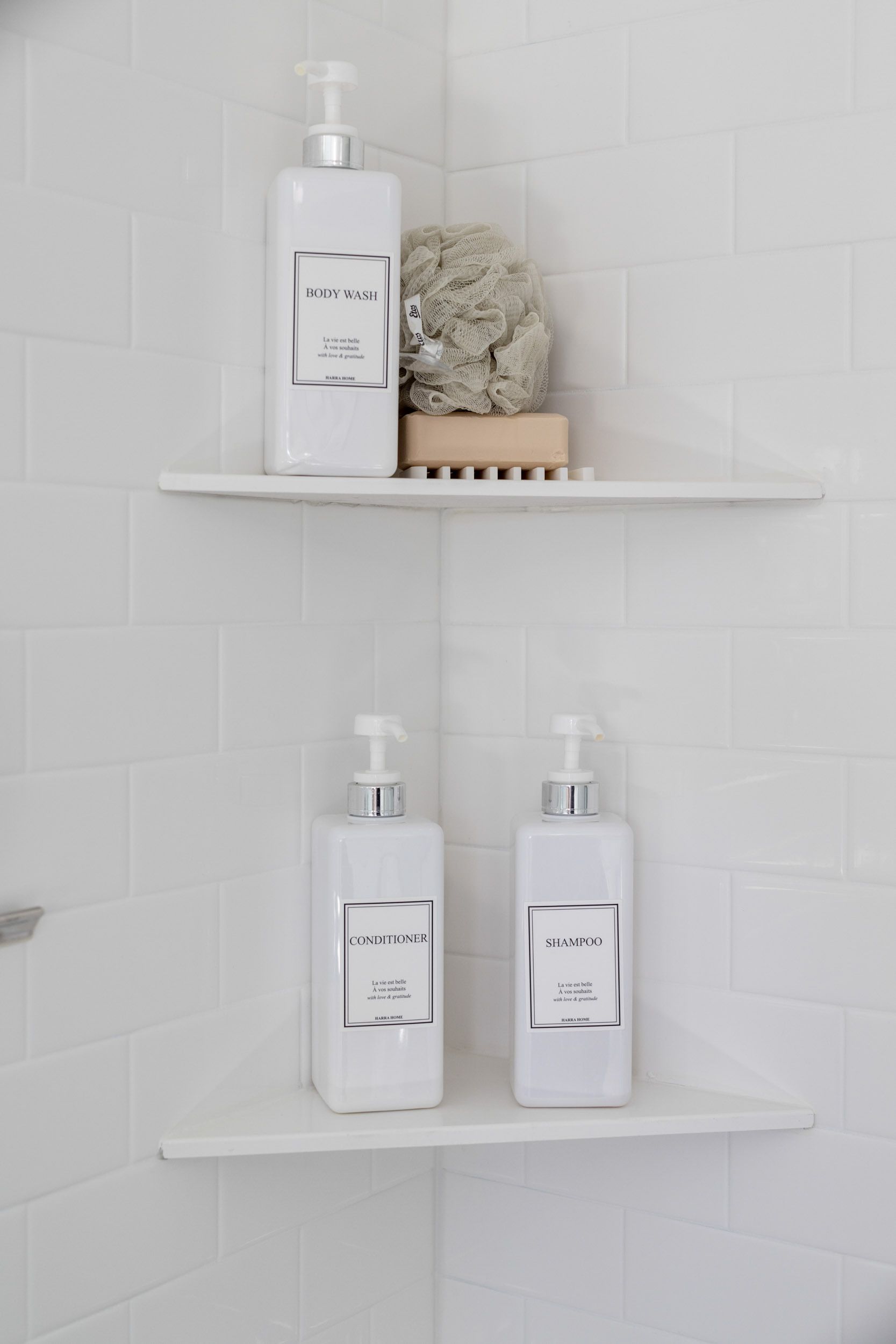 Two white corner shower shelves on white subway tile containing three matching white pump bottles and a sponge.