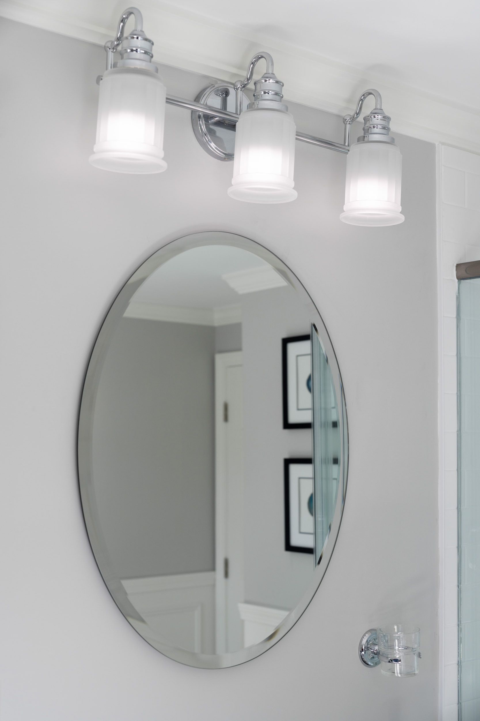 A three-light chrome vanity fixture with frosted glass shades hangs above a framed oval mirror on a light gray wall.