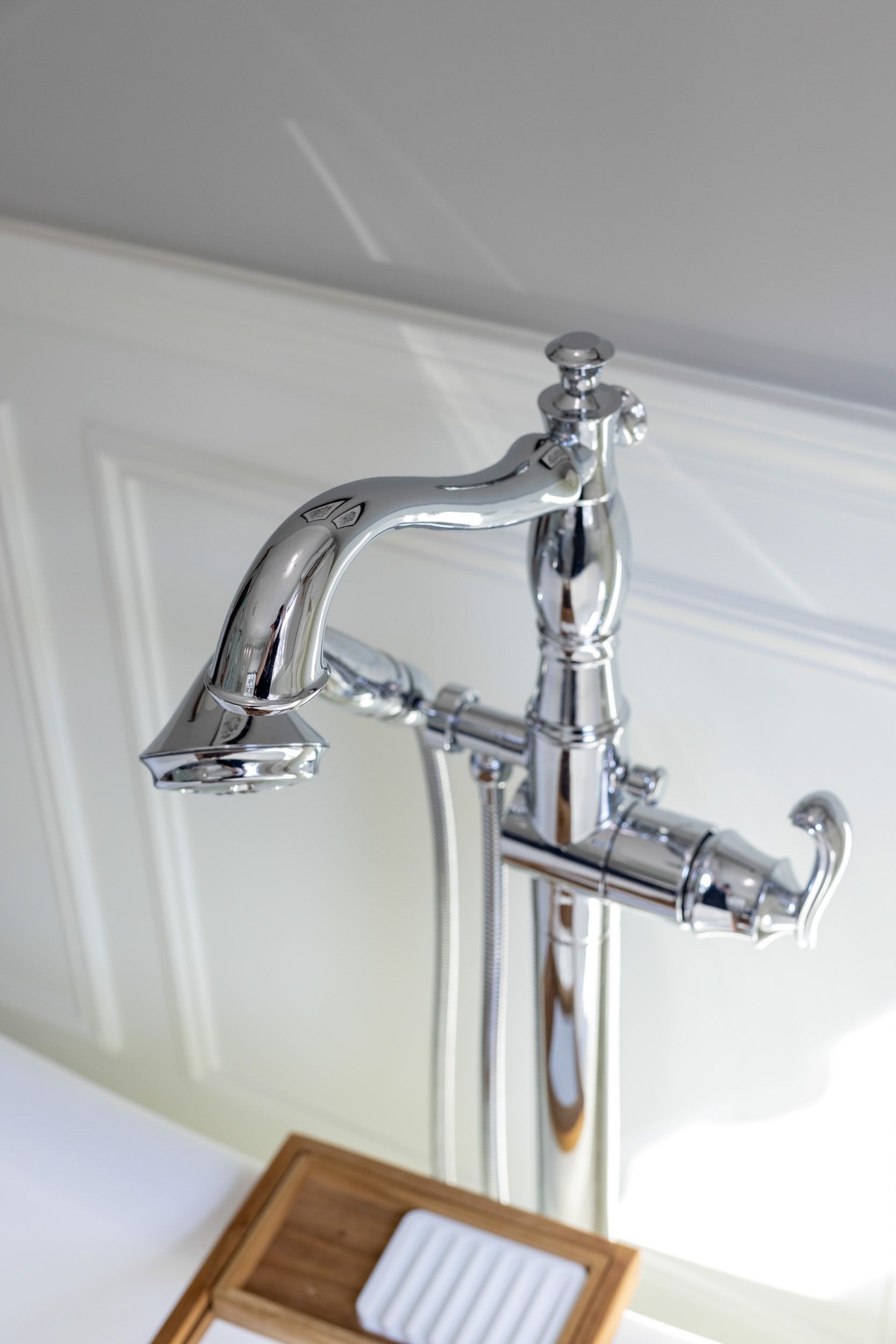 A polished chrome floor-mounted bathtub faucet with a hand shower attachment next to a wooden soap dish.
