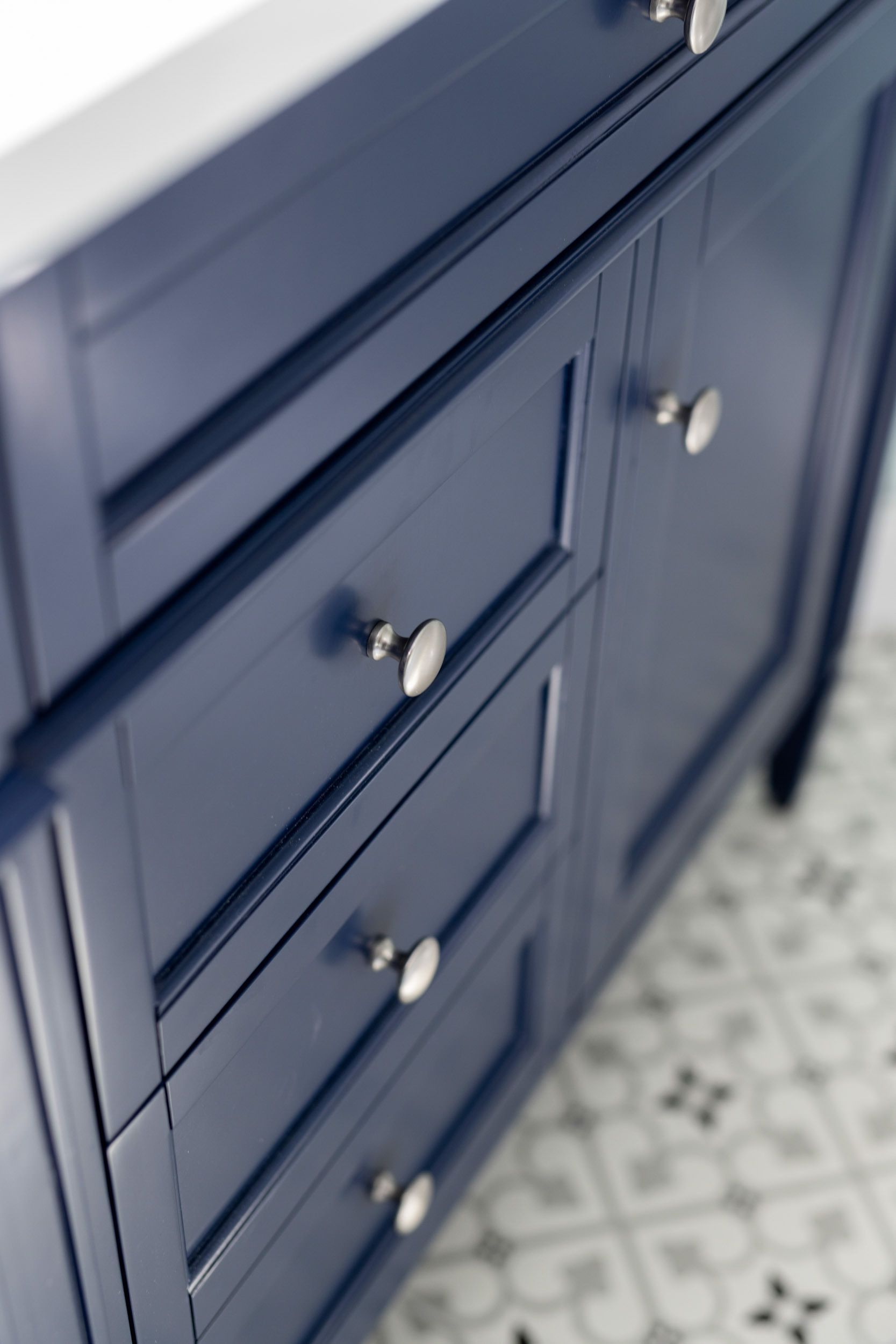 A close-up of a navy blue bathroom vanity cabinet with silver knobs, set against a patterned tile floor.