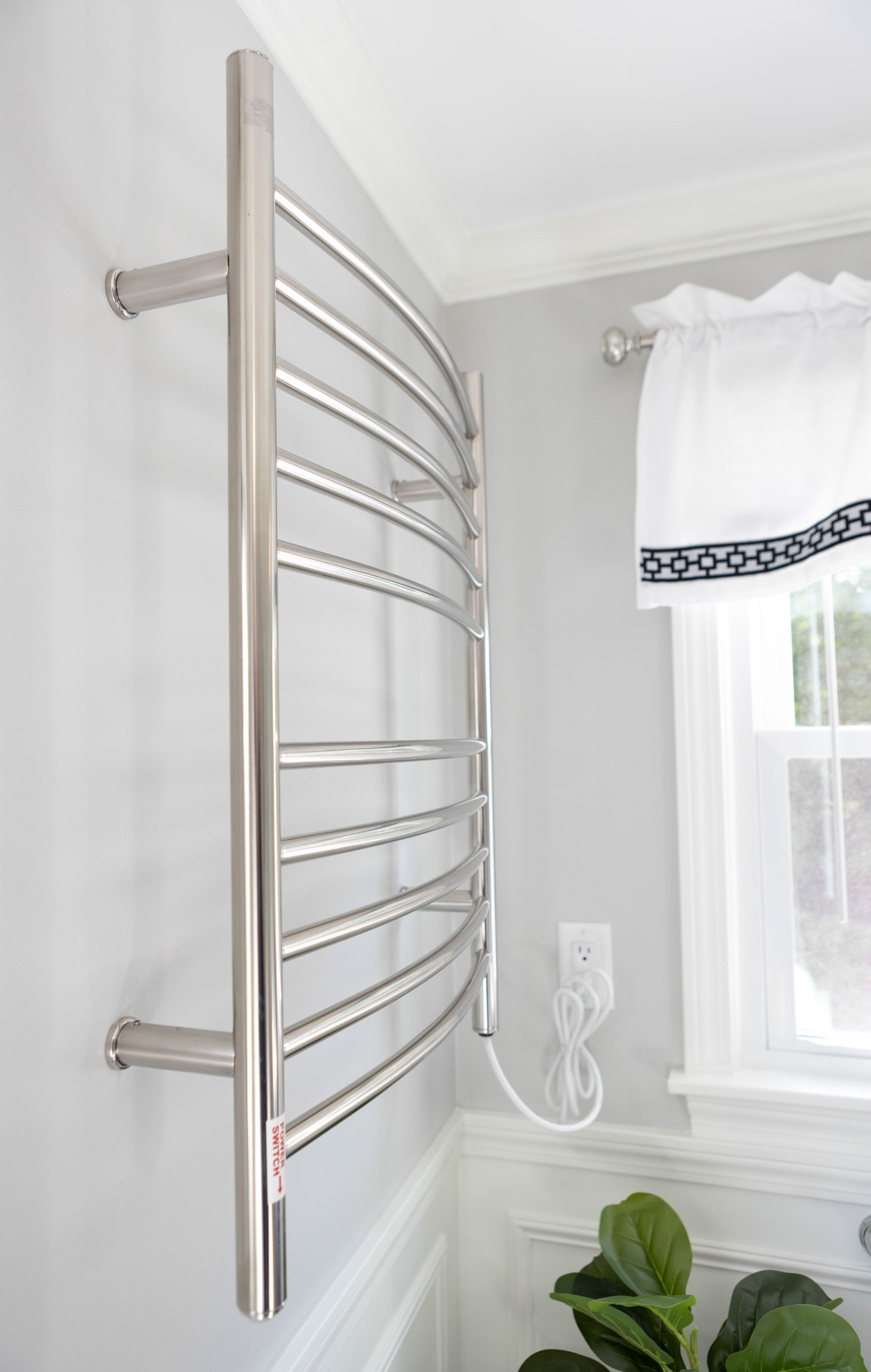 A curved, stainless steel towel warmer mounted on a grey wall next to a window with a white curtain.