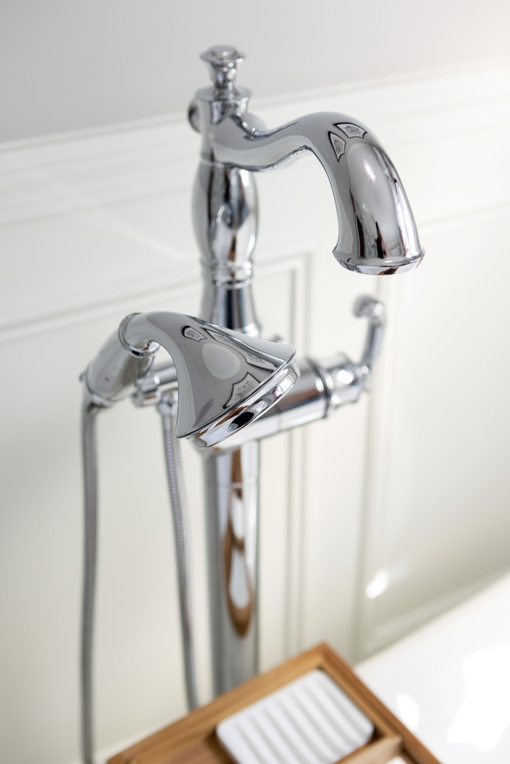 A chrome freestanding bathtub faucet and hand shower sprayer next to a wooden tray with a white soap dish.