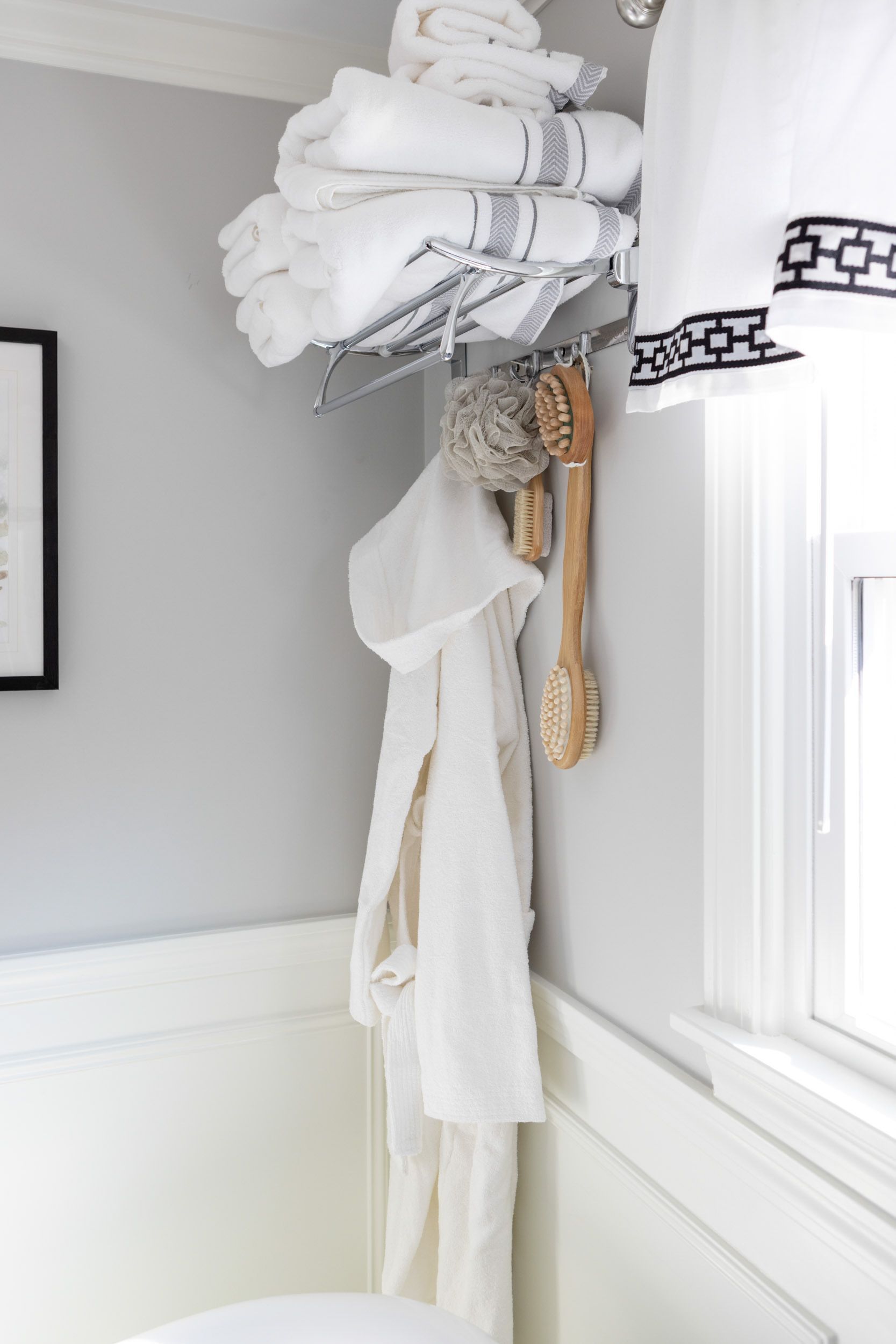 A chrome rack holding white towels above a hanging white bathrobe and wooden bath brushes on a grey bathroom wall.