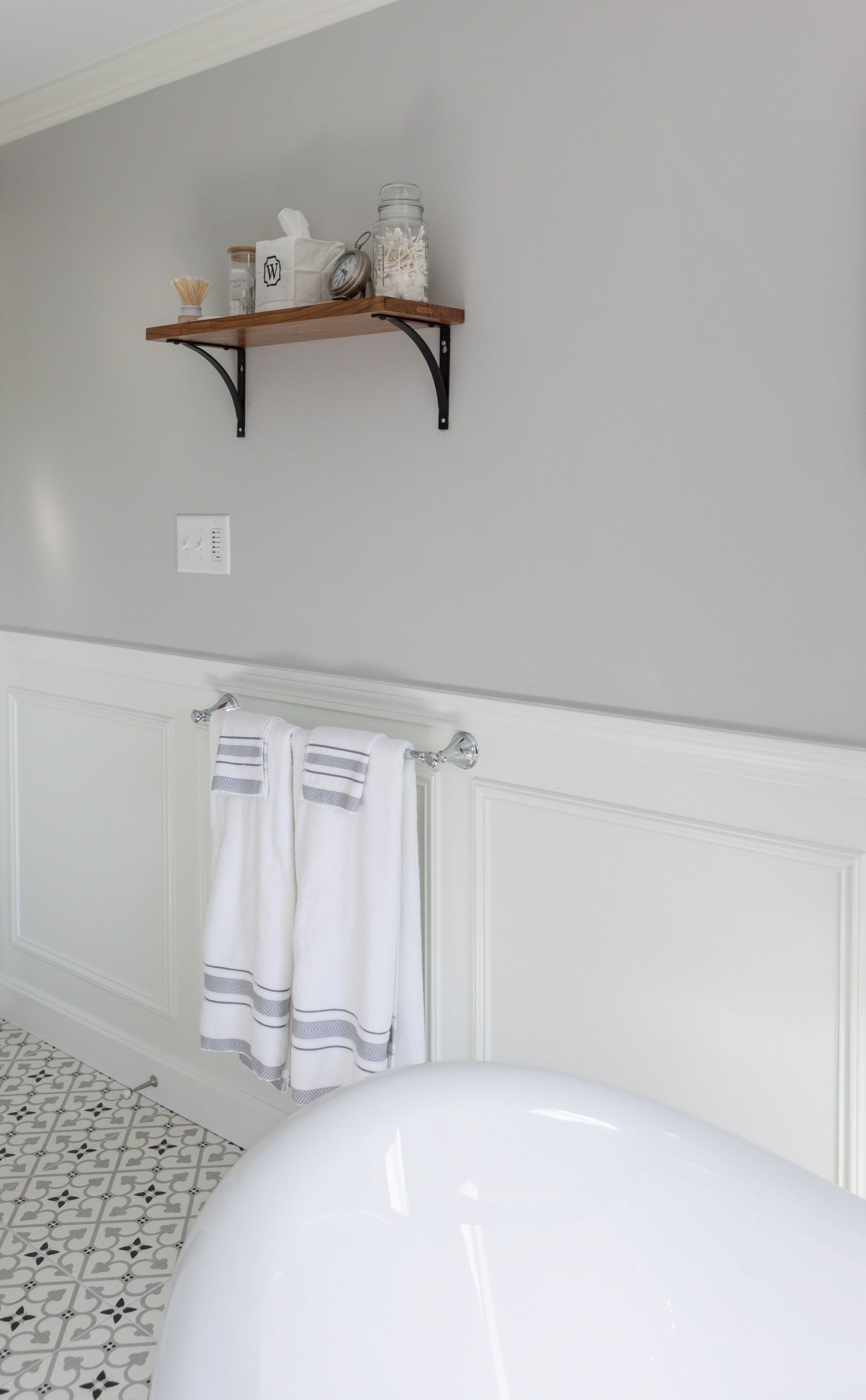 A wooden shelf with toiletries and towels hangs above a white-paneled wall, near a white bathtub in a tiled bathroom.