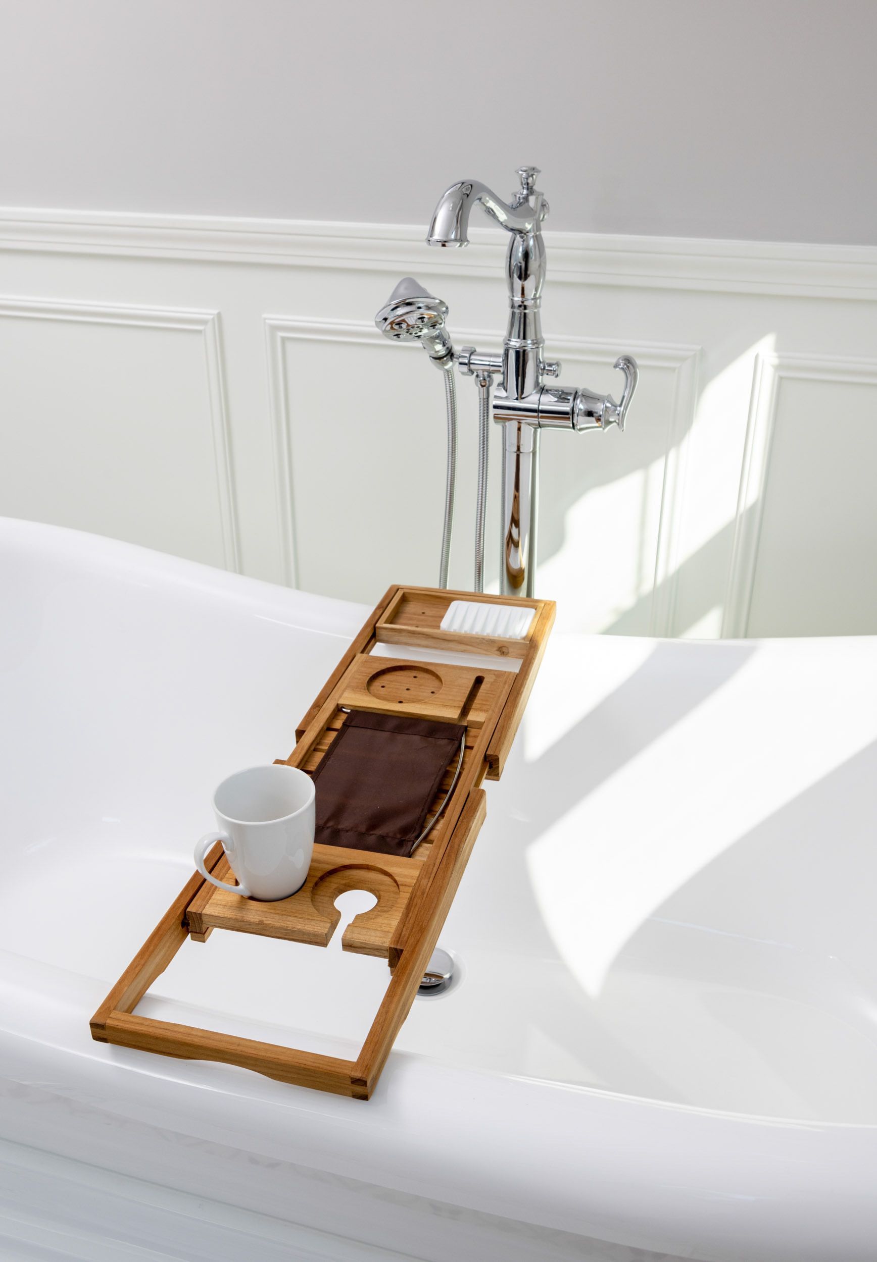A wooden bathtub caddy rests across a white porcelain tub, holding a white mug and a brown cloth near a chrome faucet.
