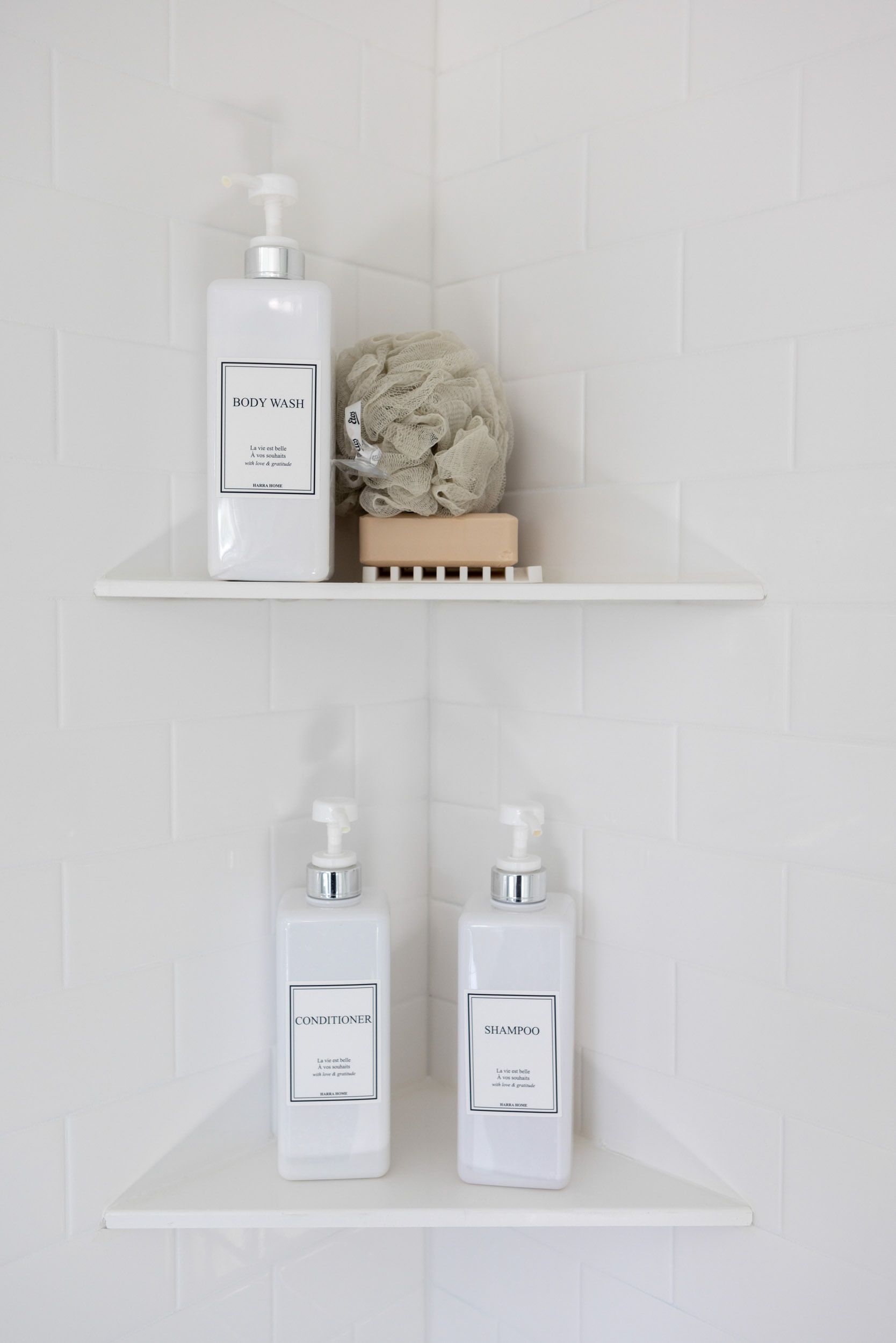 Two white tiered corner shelves in a tiled shower hold three matching pump bottles, a bath sponge, and a wooden soap dish.