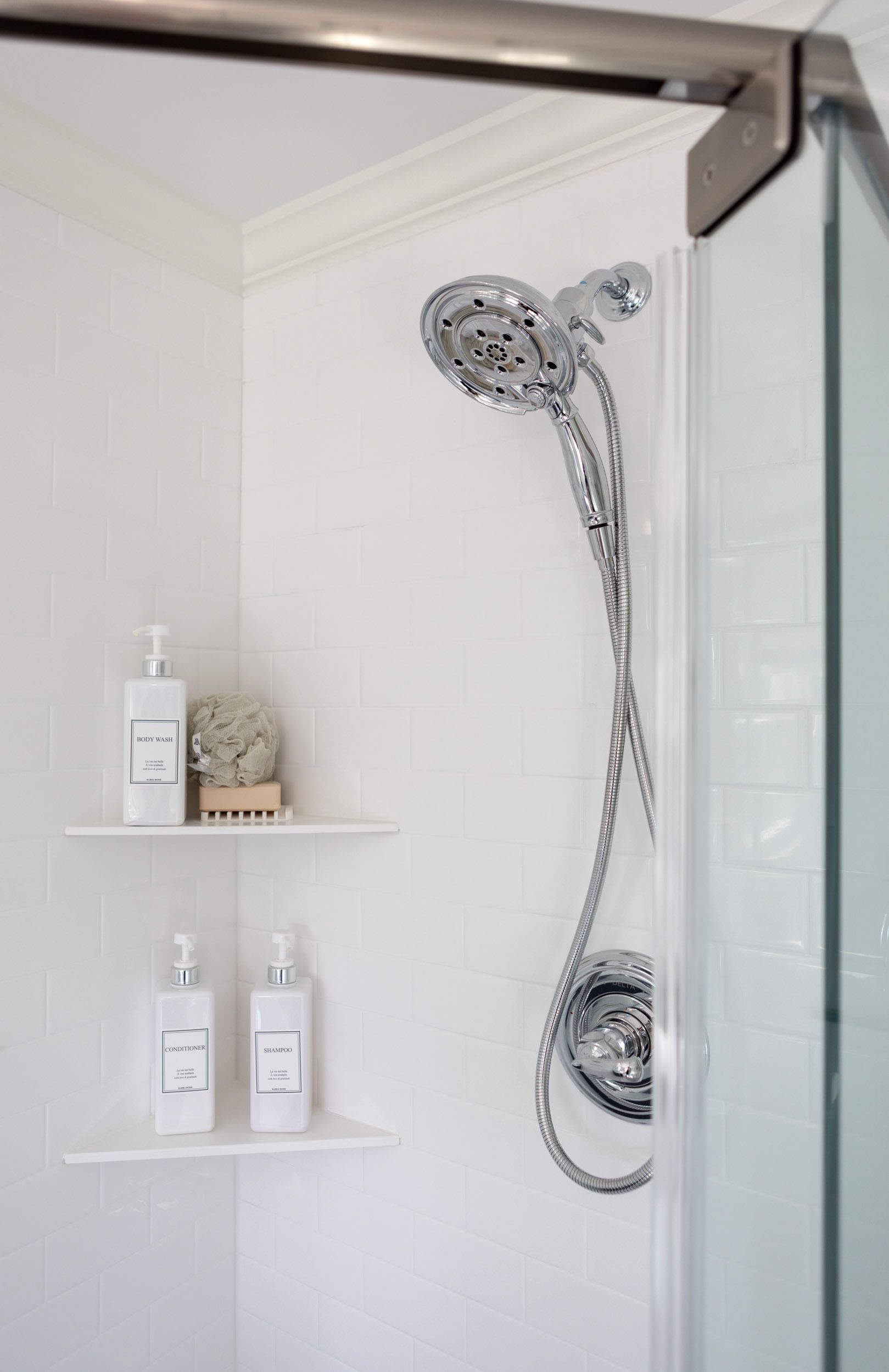 A white-tiled shower interior featuring a chrome handheld shower head, a glass door, and two white corner shelves with soap.