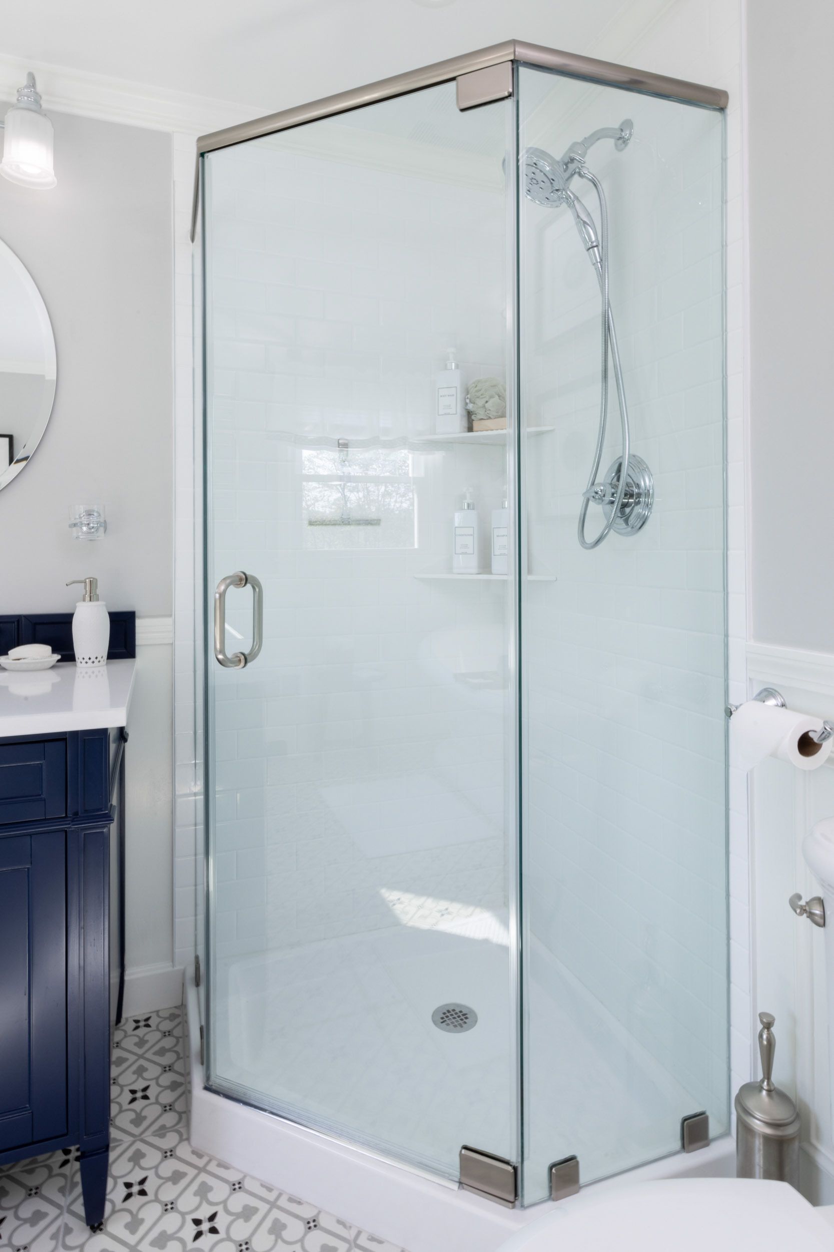 A corner glass shower stall in a modern bathroom with dark blue vanity and patterned floor tile.
