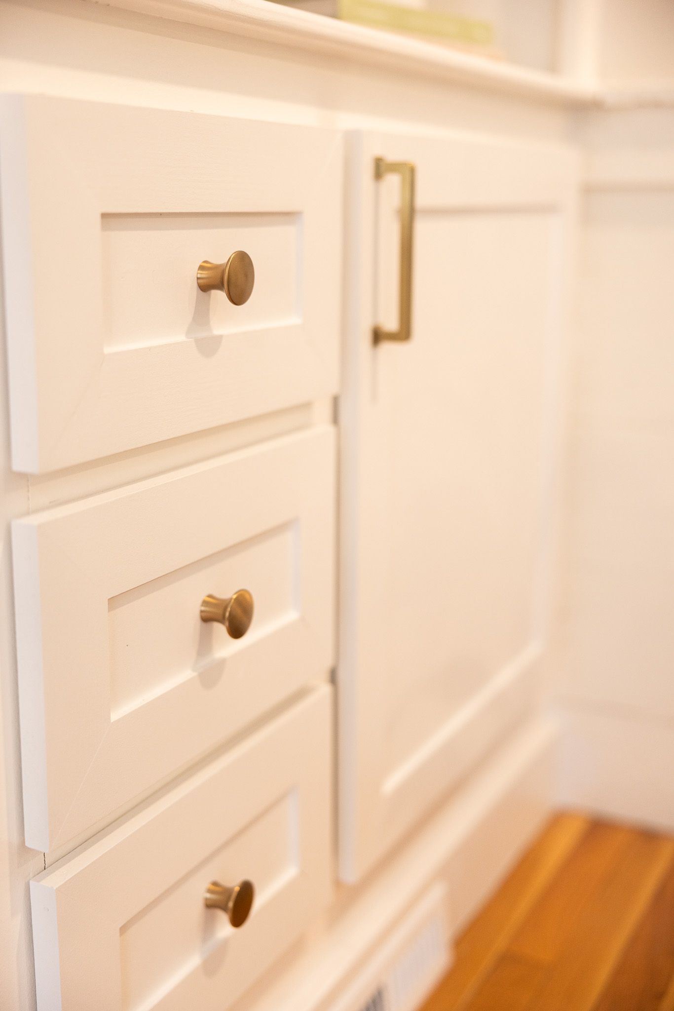 A set of white cabinets with three stacked drawers and one door, accented with round brass knobs and a brass handle.