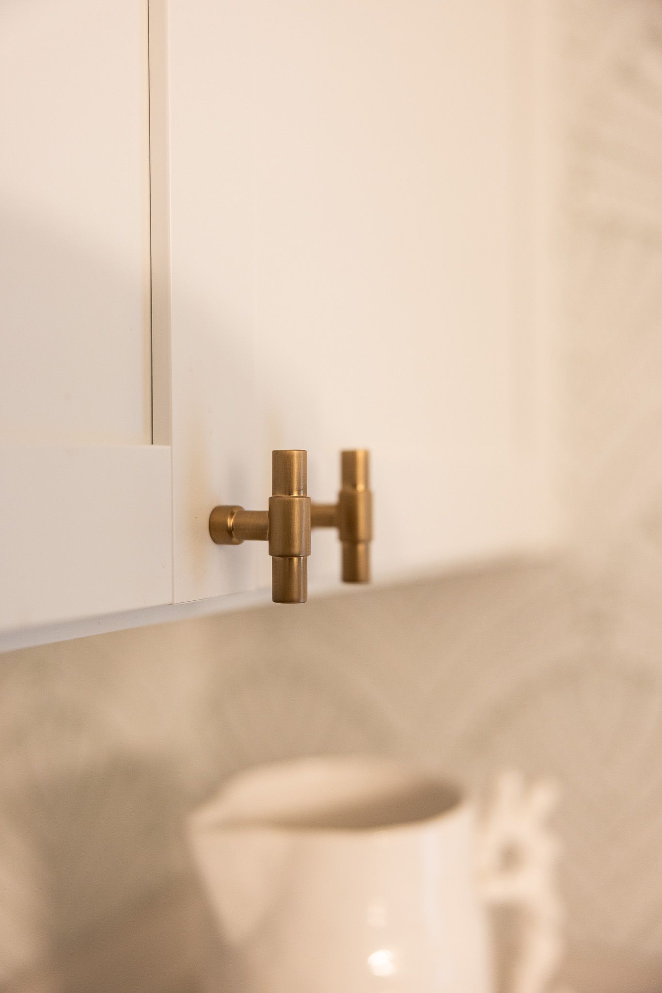 A brass T-shaped cabinet pull mounted on a white kitchen cabinet door above a ceramic pitcher.