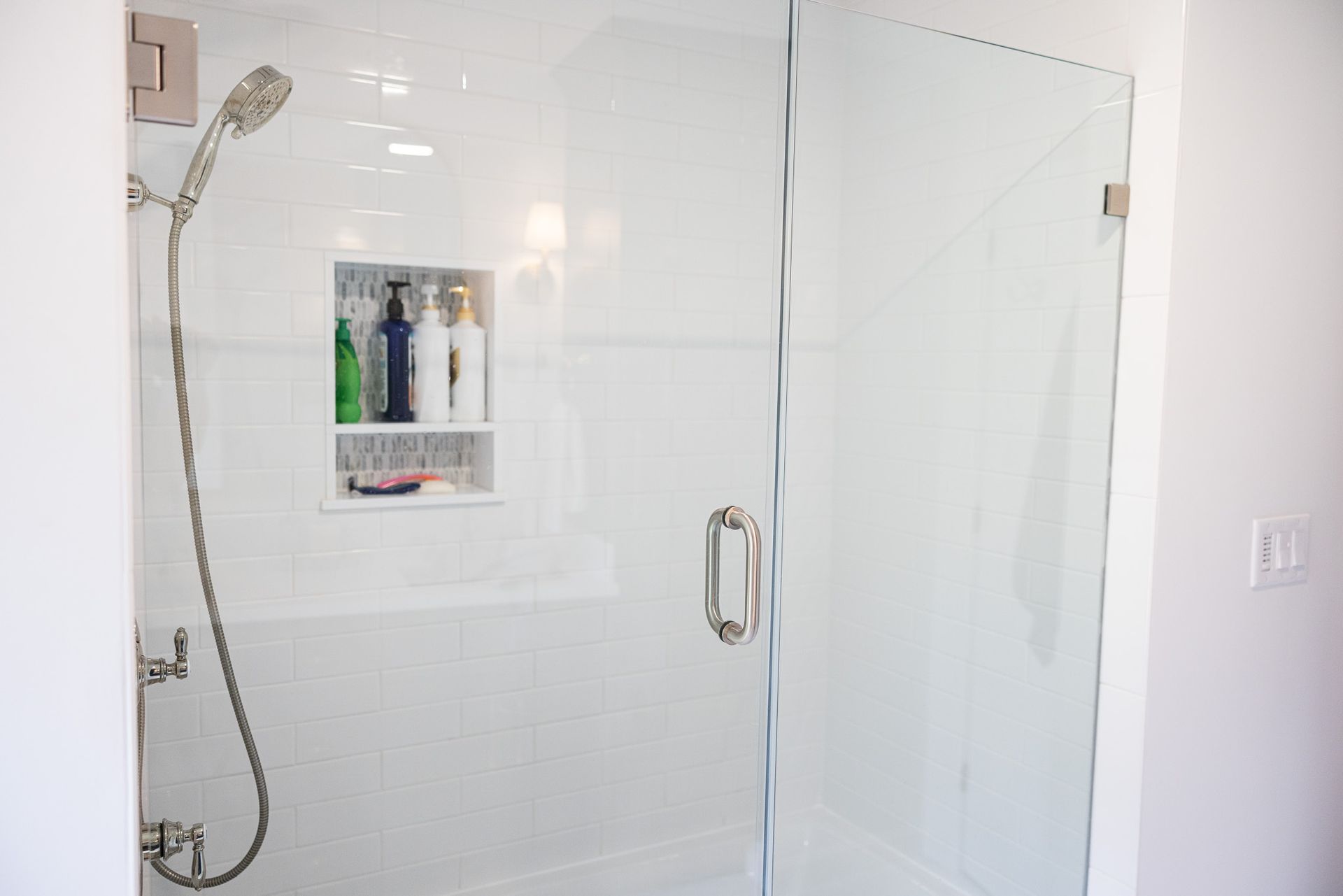 A modern walk-in shower with a glass door, a handheld showerhead, and a recessed wall niche holding several bottles.