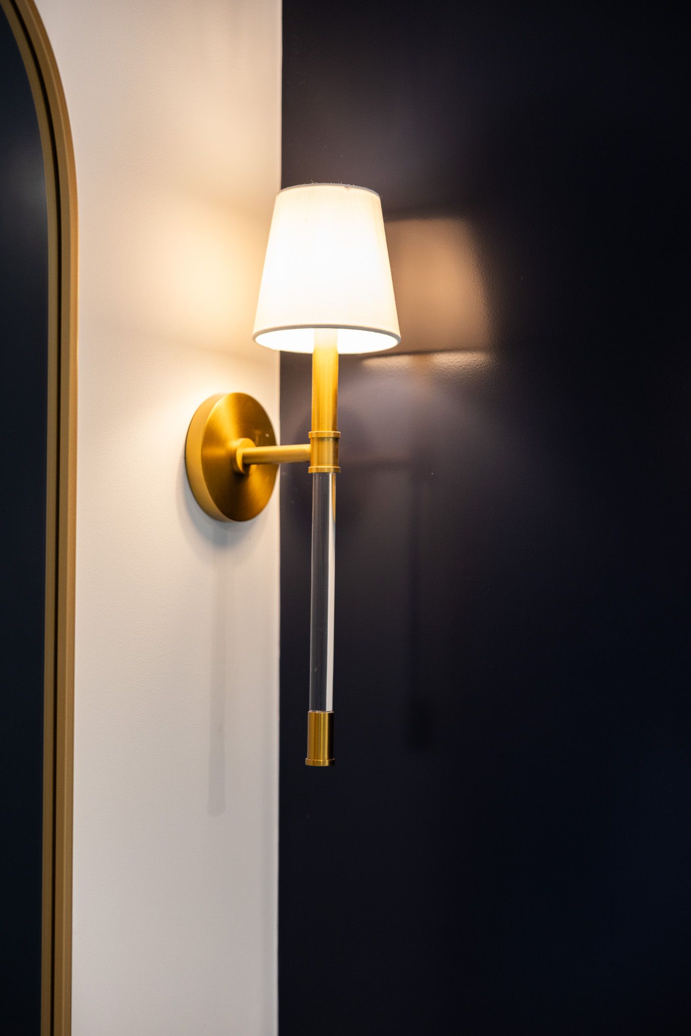 A brass wall sconce with a white fabric shade and a clear acrylic stem, mounted on a corner between a white and navy wall.