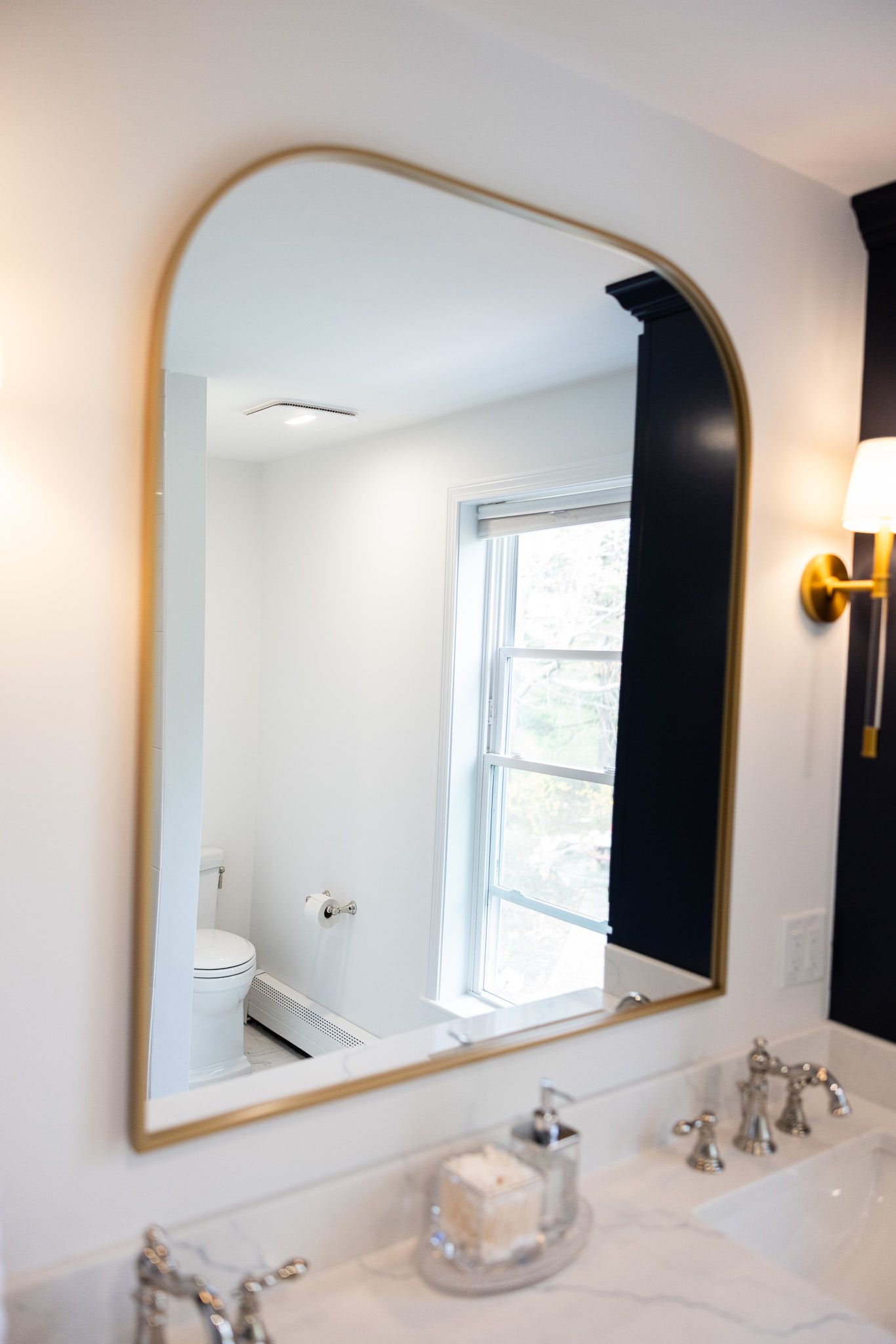 An arched gold-framed mirror hangs above a marble bathroom vanity with a faucet, soap dish, and a reflection of a window.
