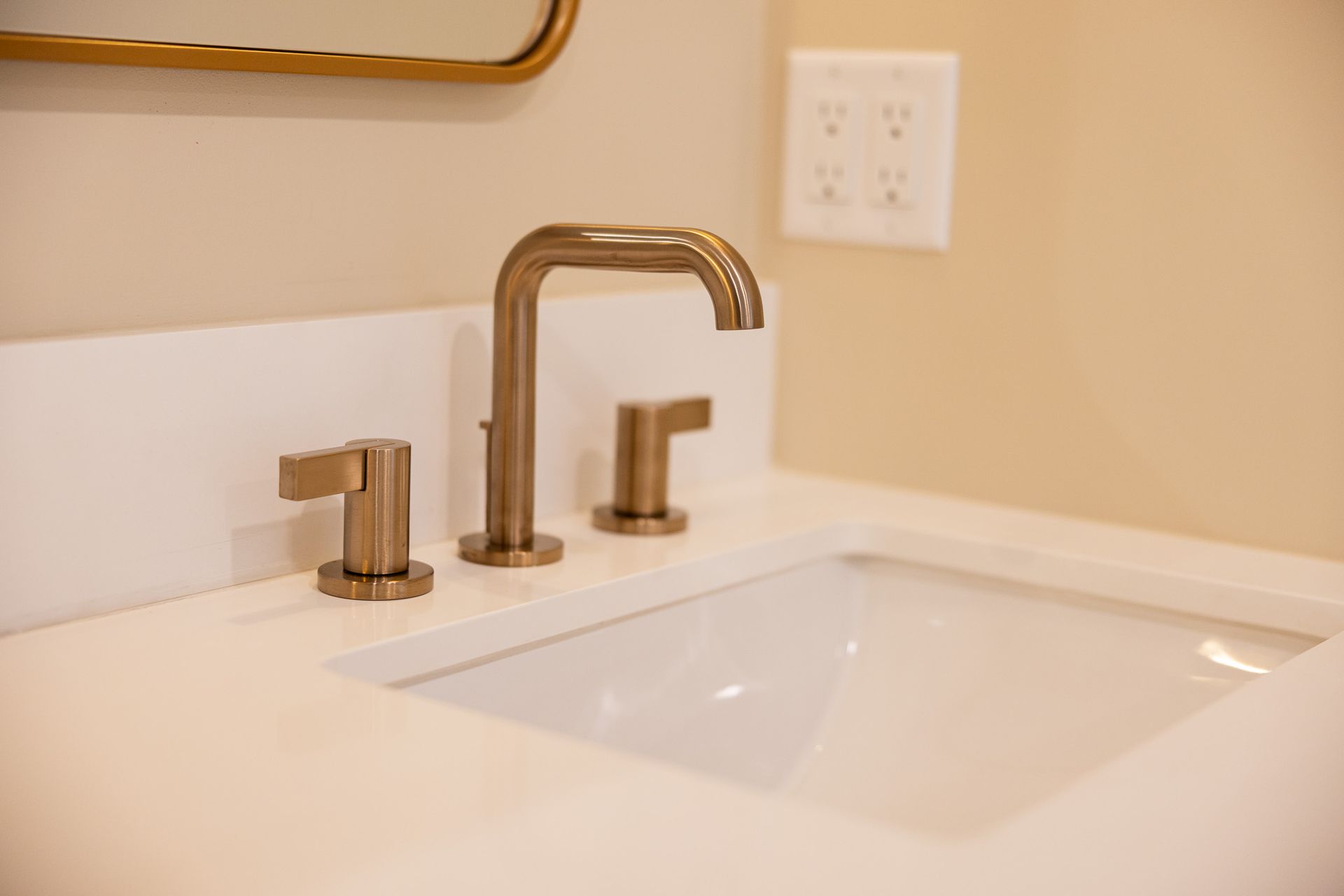A modern, brushed-gold faucet with two handles centered over a white rectangular bathroom sink against a beige wall.