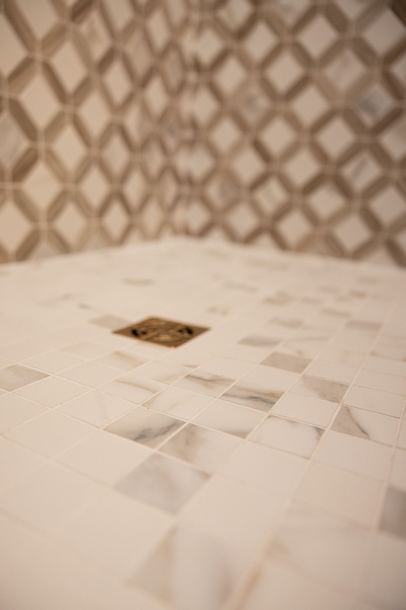White and grey marble tiled shower floor with a square bronze drain, set against patterned geometric wall tiles.