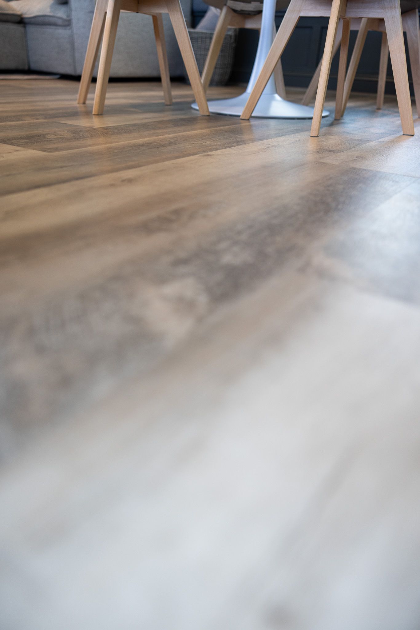 A low-angle view of wood-look flooring with blurred chair legs and a dining table base in the background.