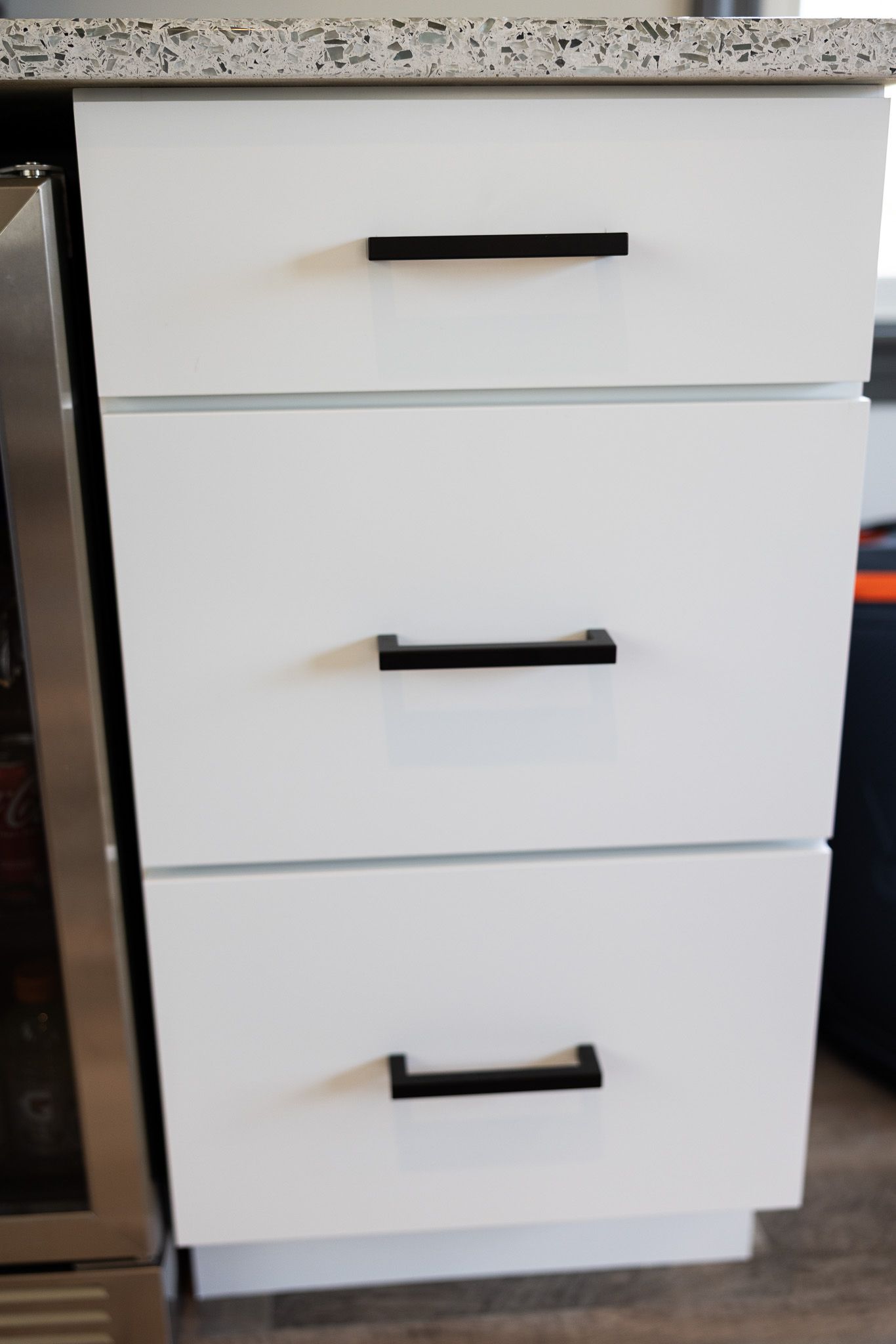 A stack of three white kitchen drawers with black rectangular handles set under a speckled granite countertop.