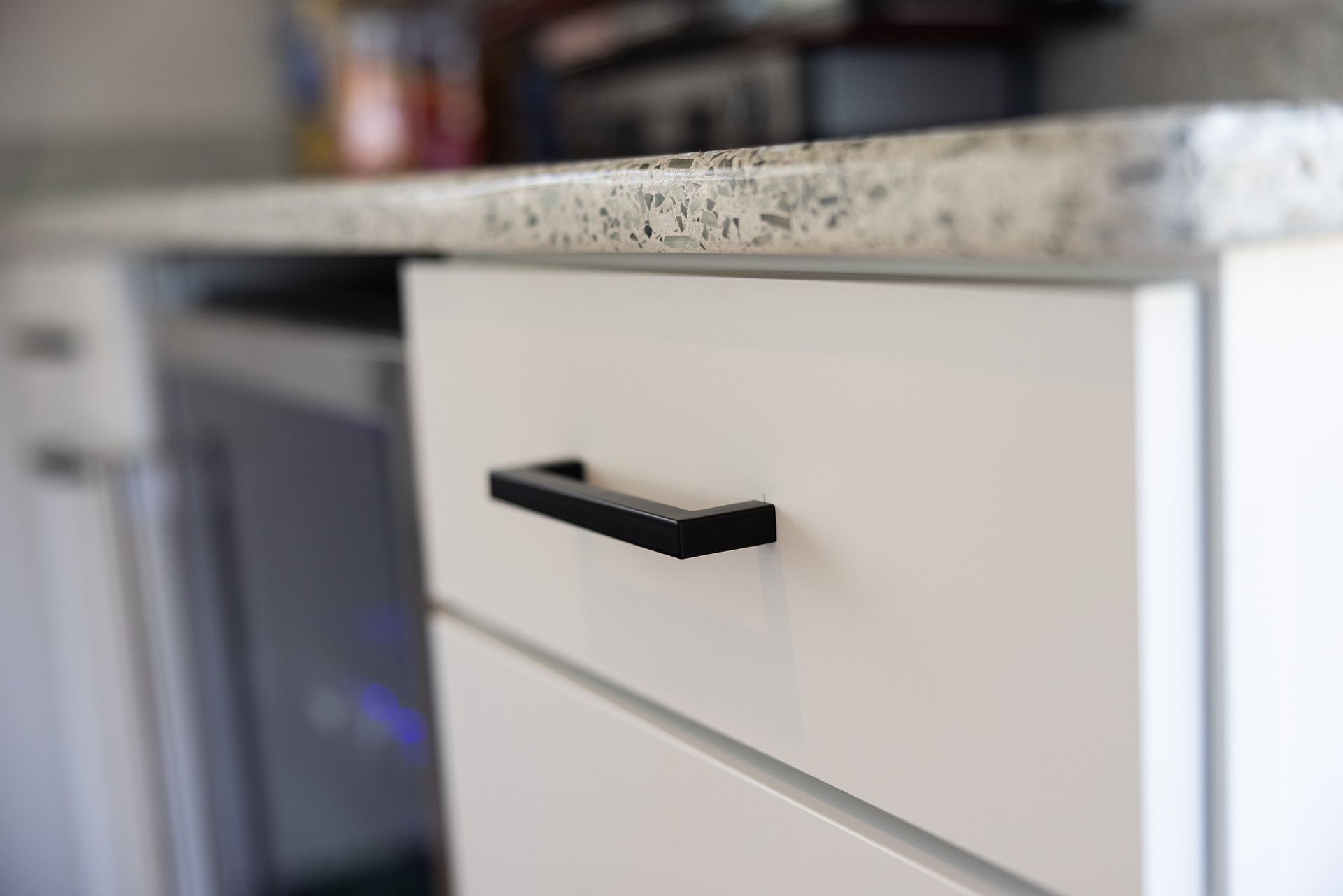 A white kitchen cabinet drawer with a black handle, positioned beneath a speckled granite countertop.