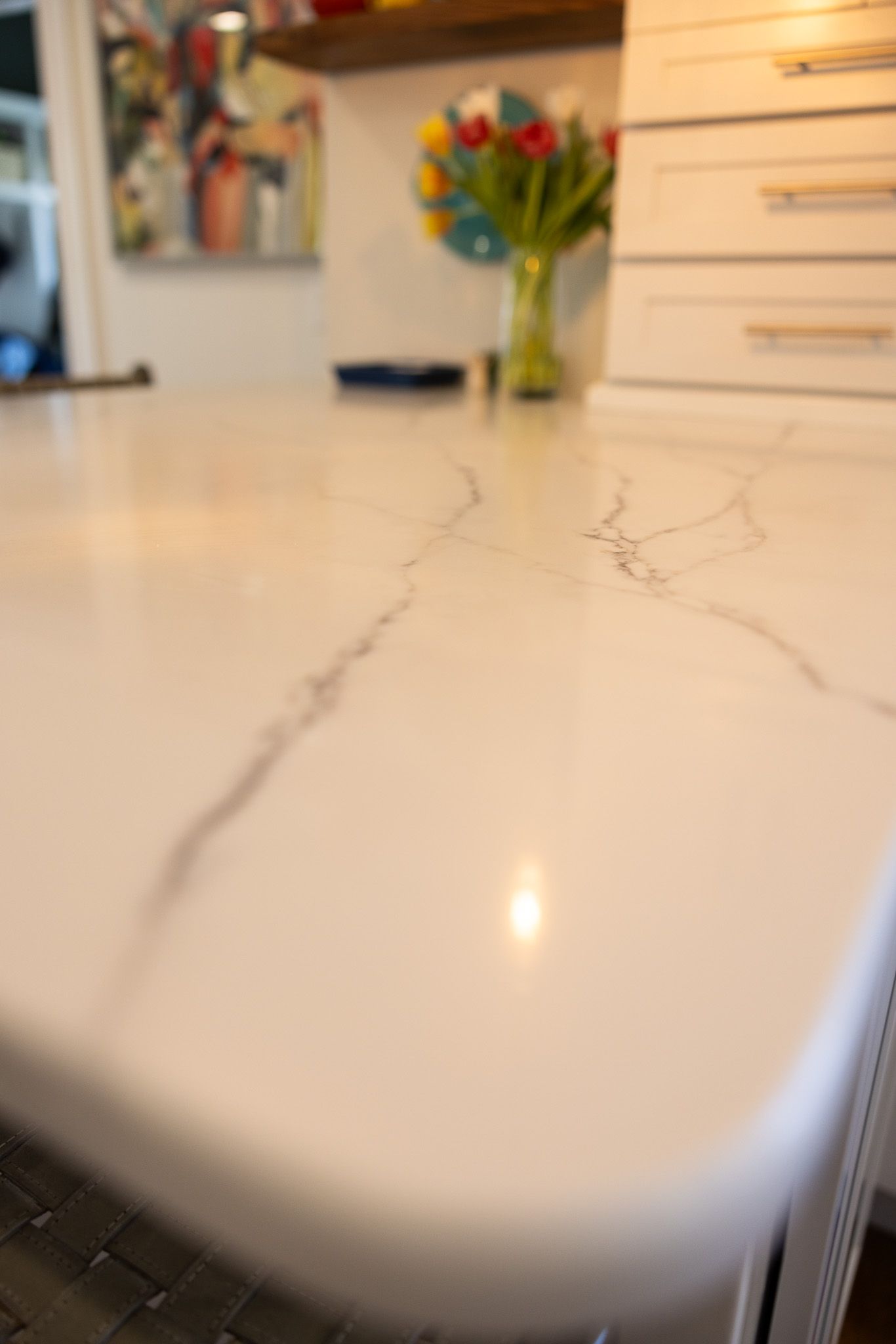 A polished white marble countertop with grey veining, angled in a bright kitchen with a vase of flowers in the background.