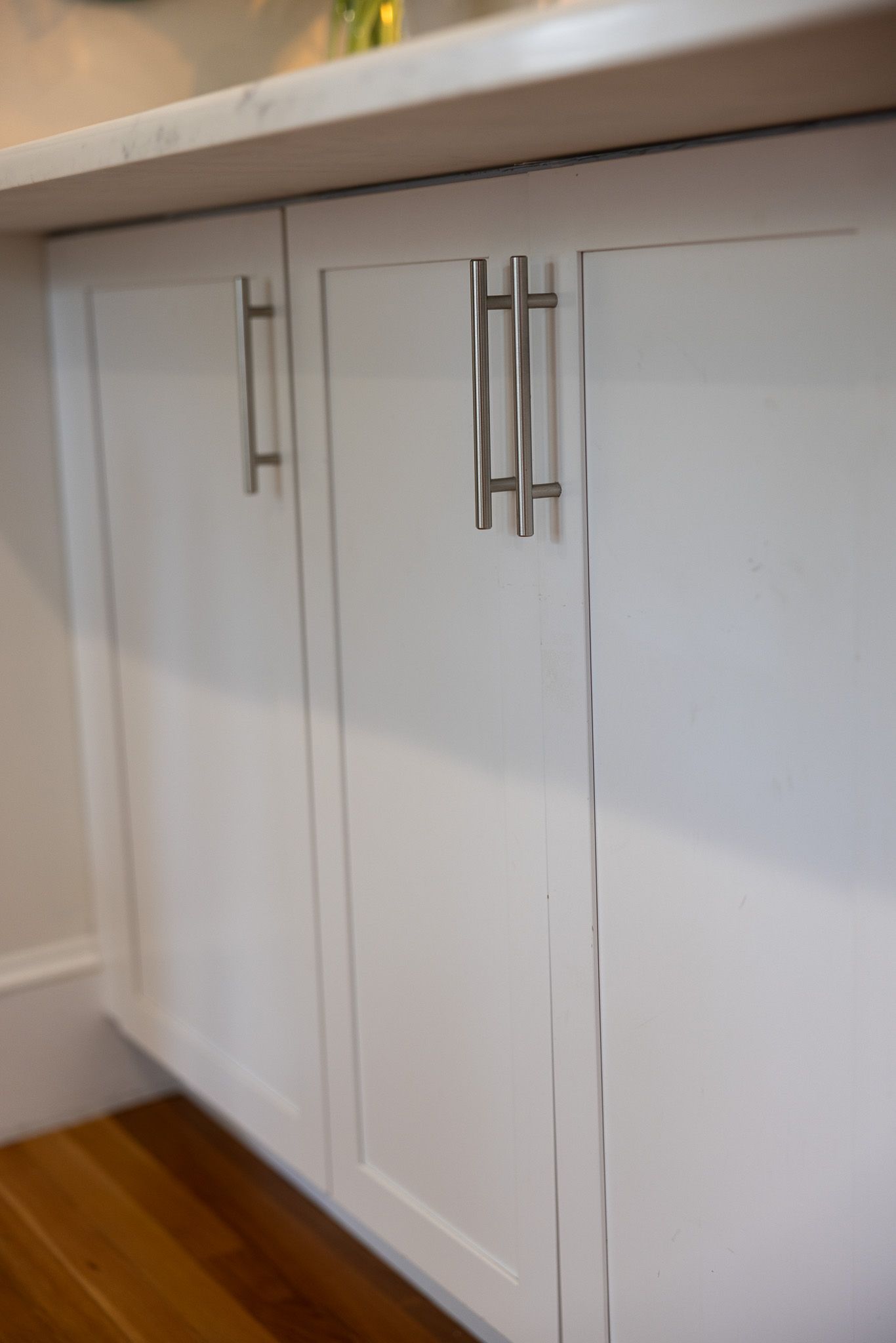 Close-up of white cabinet doors with silver bar handles underneath a countertop, positioned above a hardwood floor.