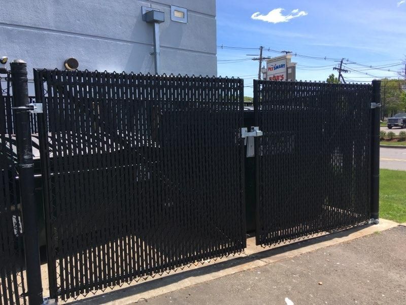 A black double-gate chain-link fence with privacy slats installed outside a building on a sunny day.