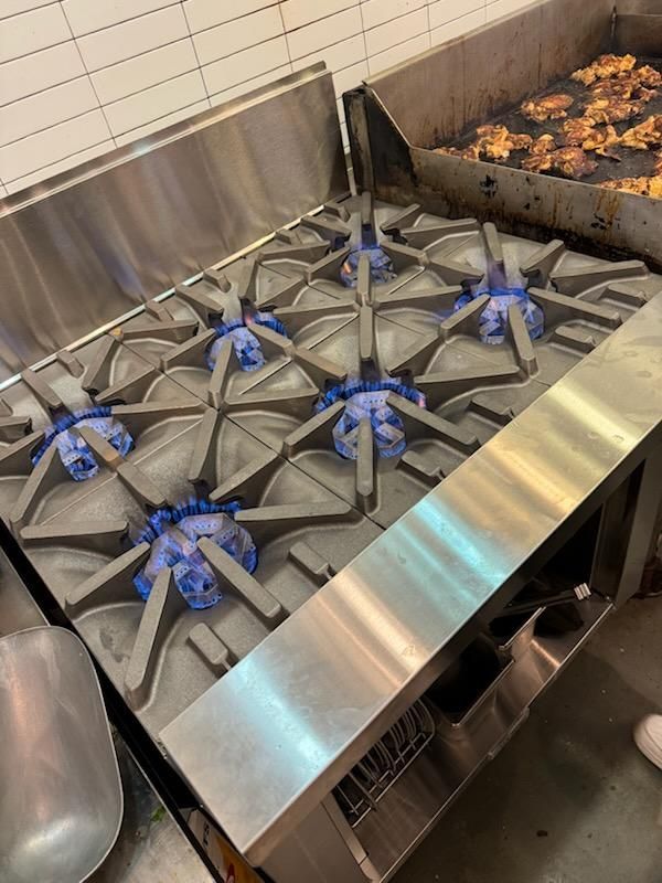 A commercial kitchen stove with four lit blue gas burners, situated next to a flat-top grill cooking pieces of chicken.