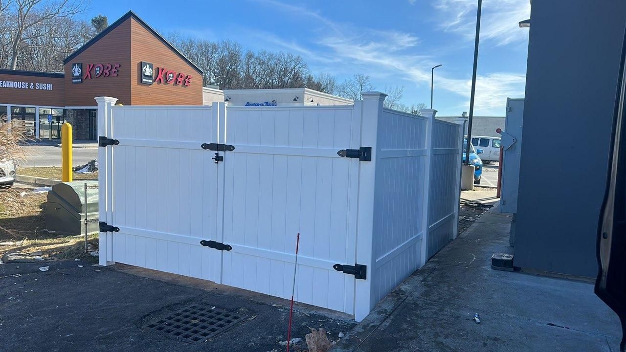 A white vinyl privacy fence enclosure for a dumpster stands in a paved lot in front of a shopping center.
