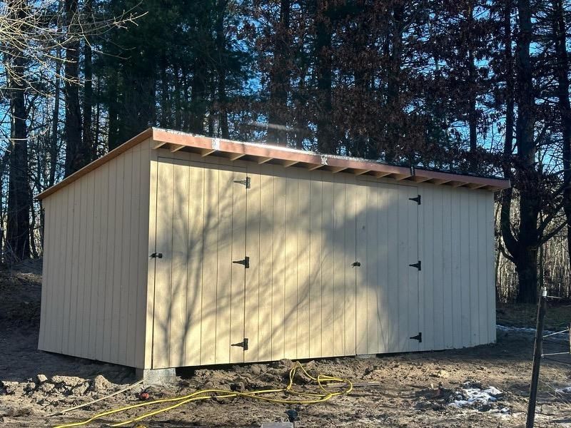 A tan, rectangular wooden shed with double doors sits in a wooded area on a dirt clearing.