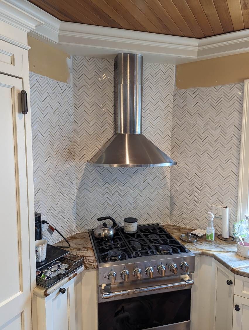 A stainless steel range hood installed in a corner over a gas stove with white herringbone-patterned tile backsplash.