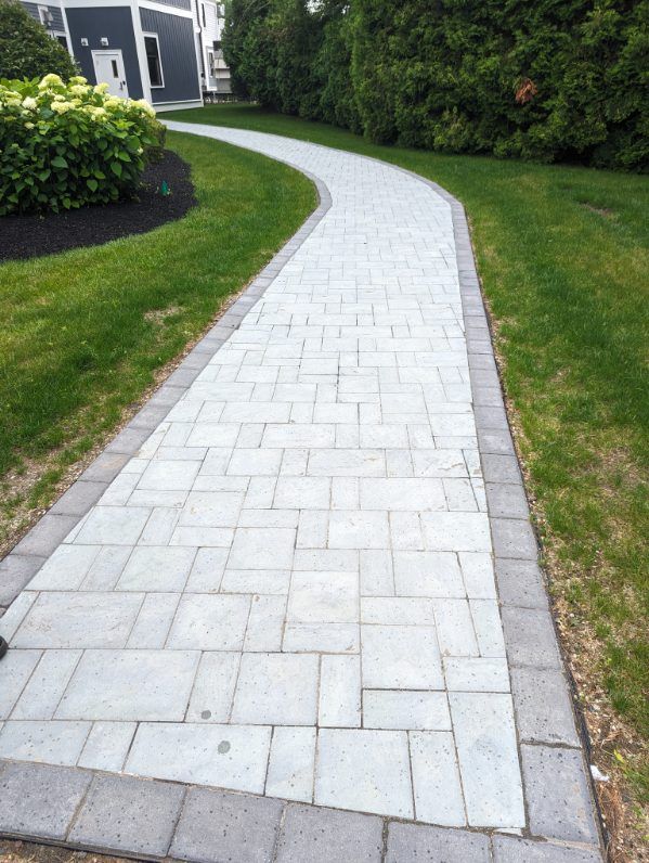 A paved stone walkway curves through a grassy yard alongside a house and a row of evergreen shrubs.