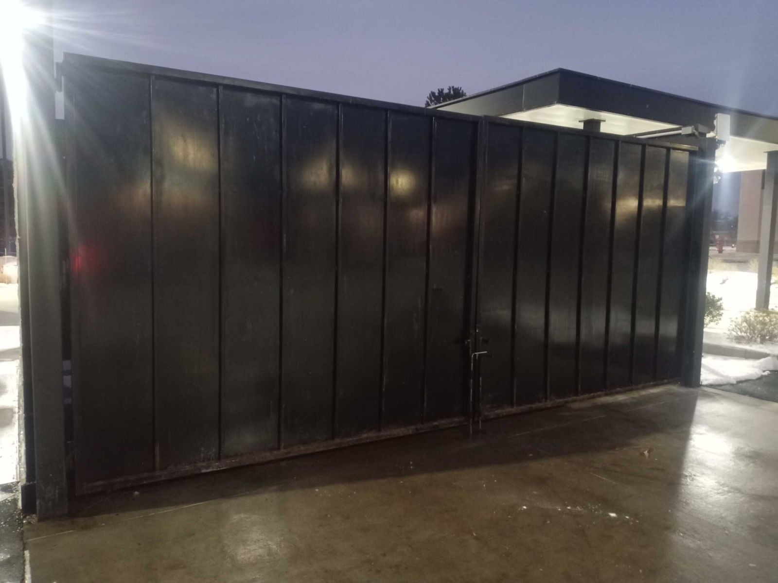 A large, dark metal gate with vertical panels set against a concrete driveway at twilight.
