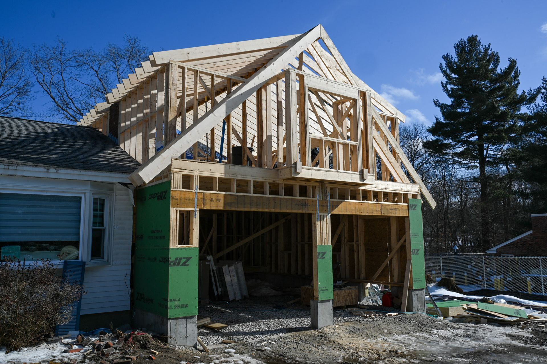 A house addition under construction with exposed wooden framing and green sheathing boards against a blue sky.