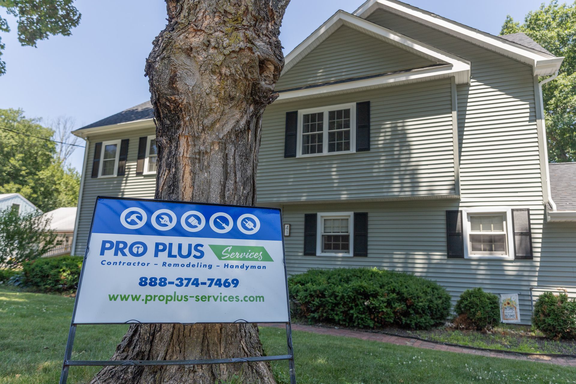A sign for Pro Plus Services stands in front of a gray two-story house next to a large tree on a sunny day.