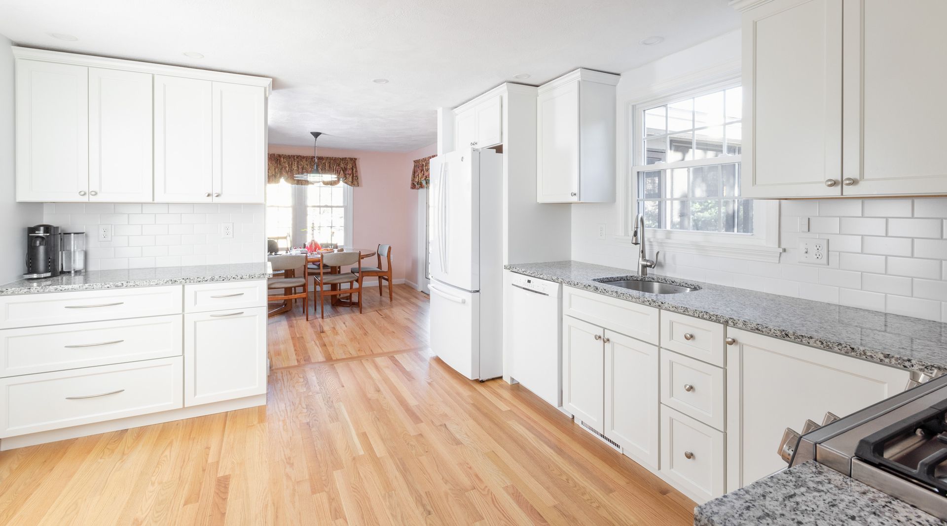 A bright, modern kitchen with white cabinets, light wood floors, gray granite countertops, and a dining area in the back.