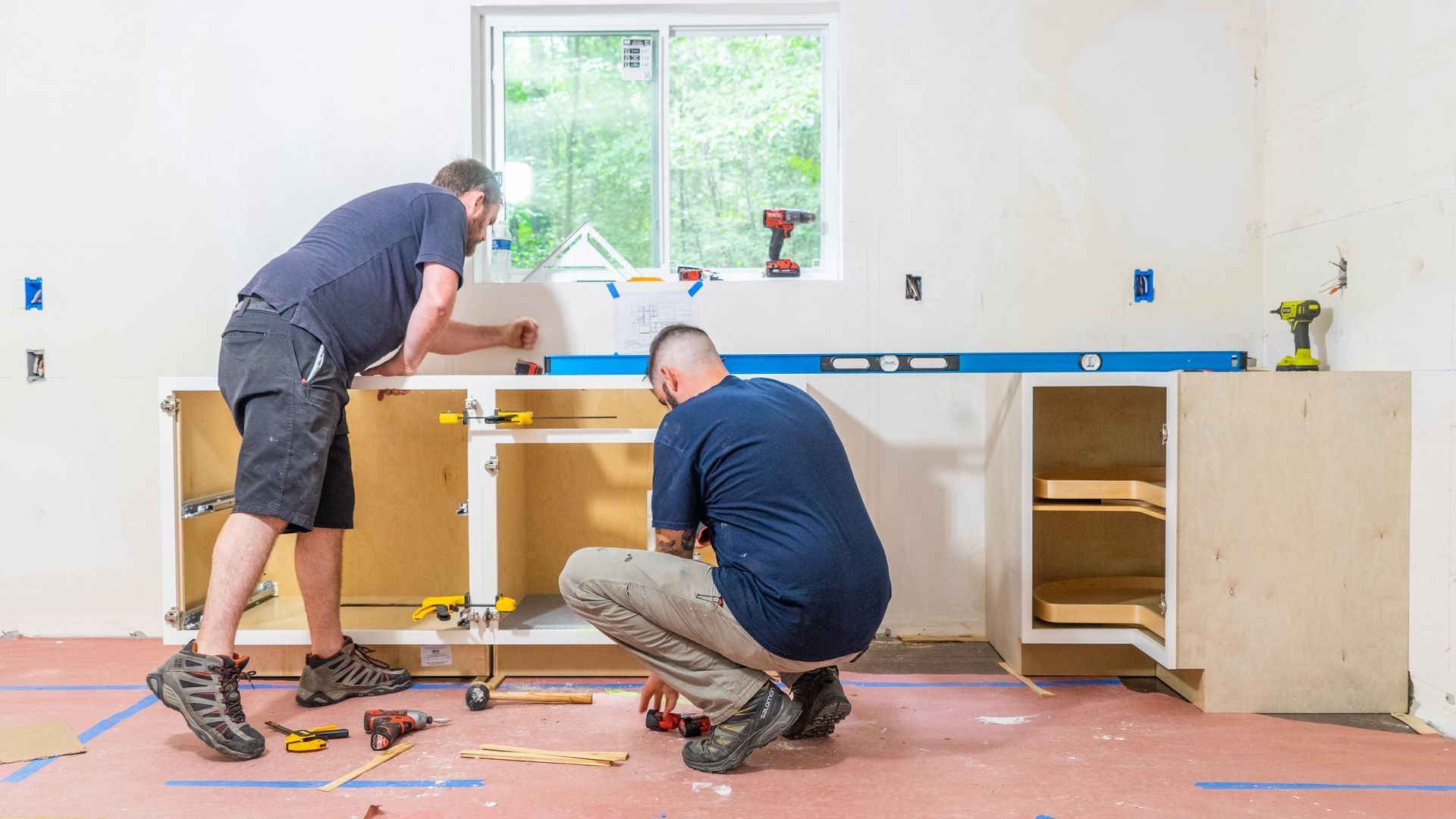 Two people in a kitchen under construction installing cabinets and using a long level along the wall.