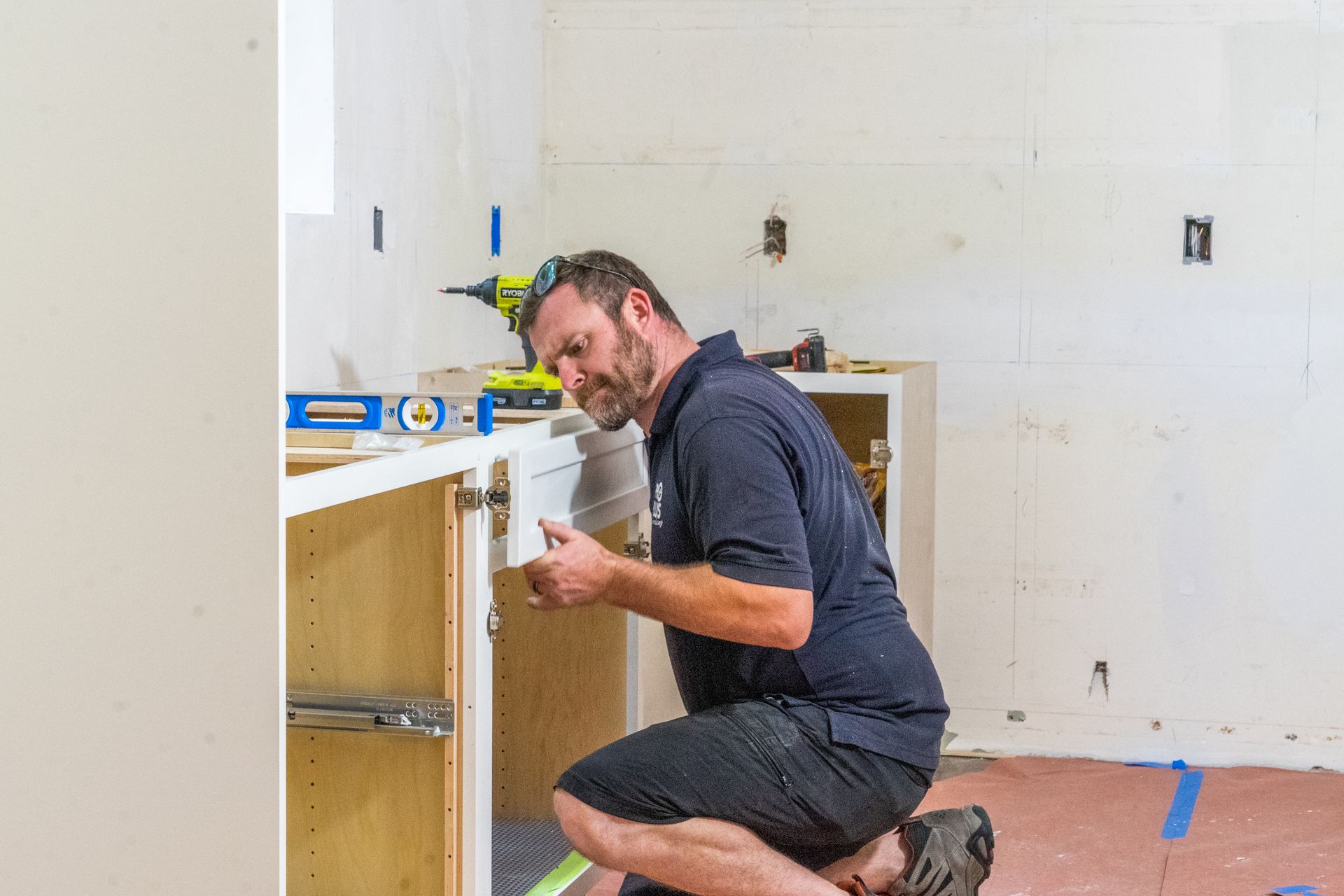 A person kneels while installing kitchen cabinets, using a level to ensure the cabinet alignment.