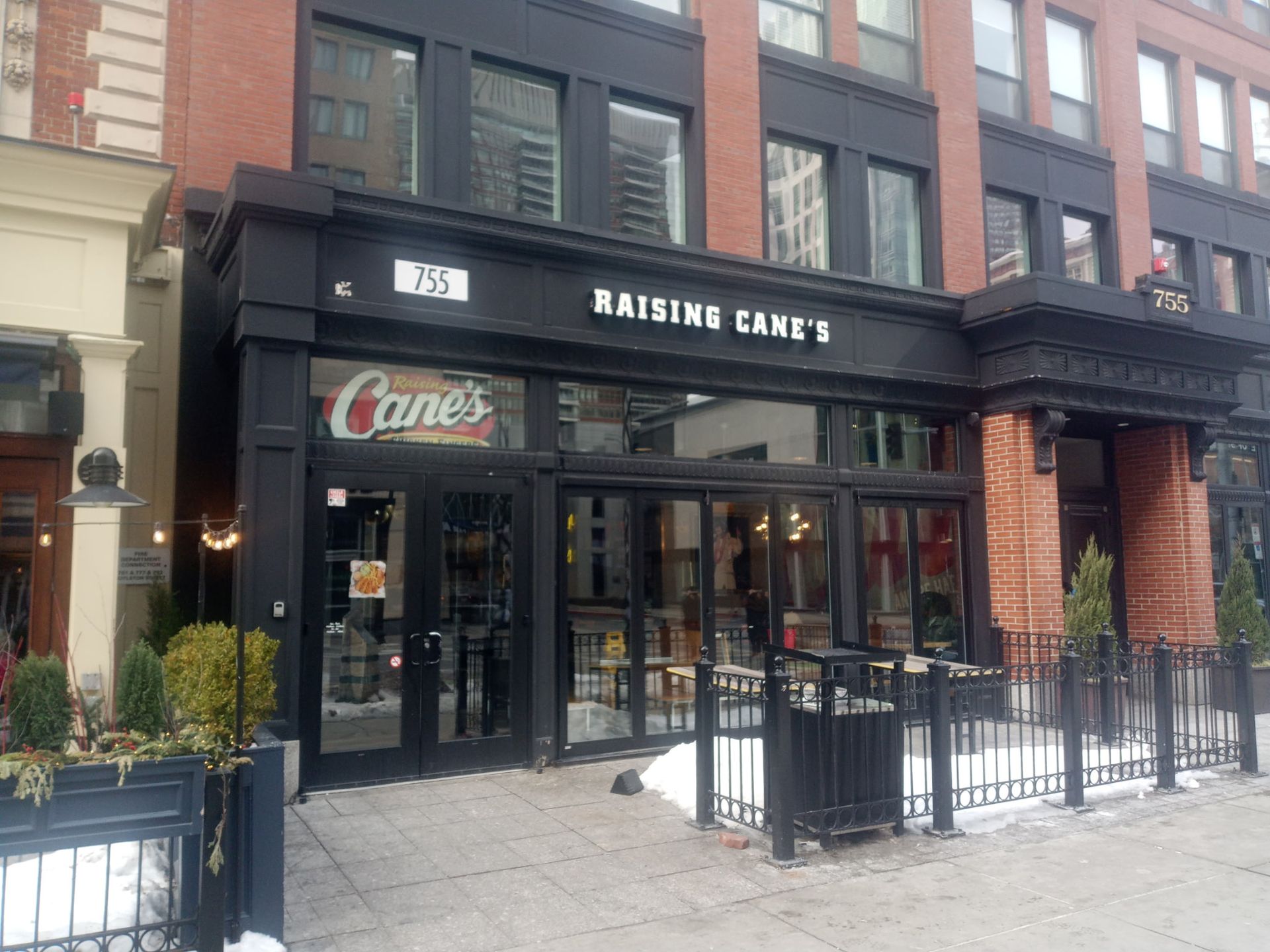 A street-level view of a Raising Cane's restaurant storefront with a black facade and large glass windows on a city street.