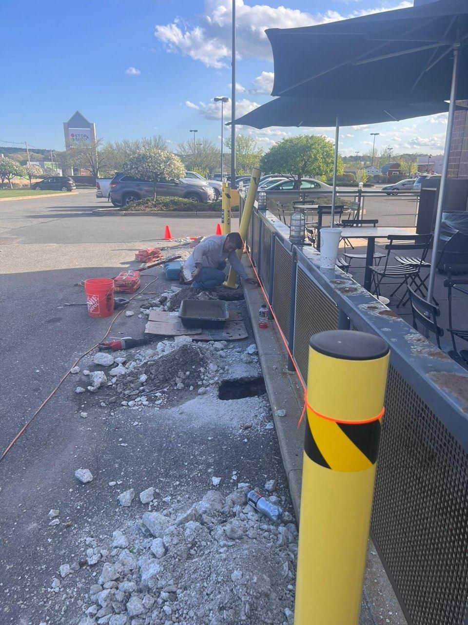 A worker cuts concrete next to a restaurant patio, with safety cones and debris on the ground in a parking lot.