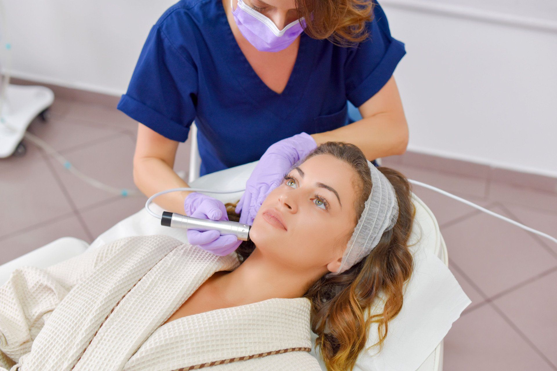 Woman receiving a facial treatment in a salon; a technician holds a device over her face.