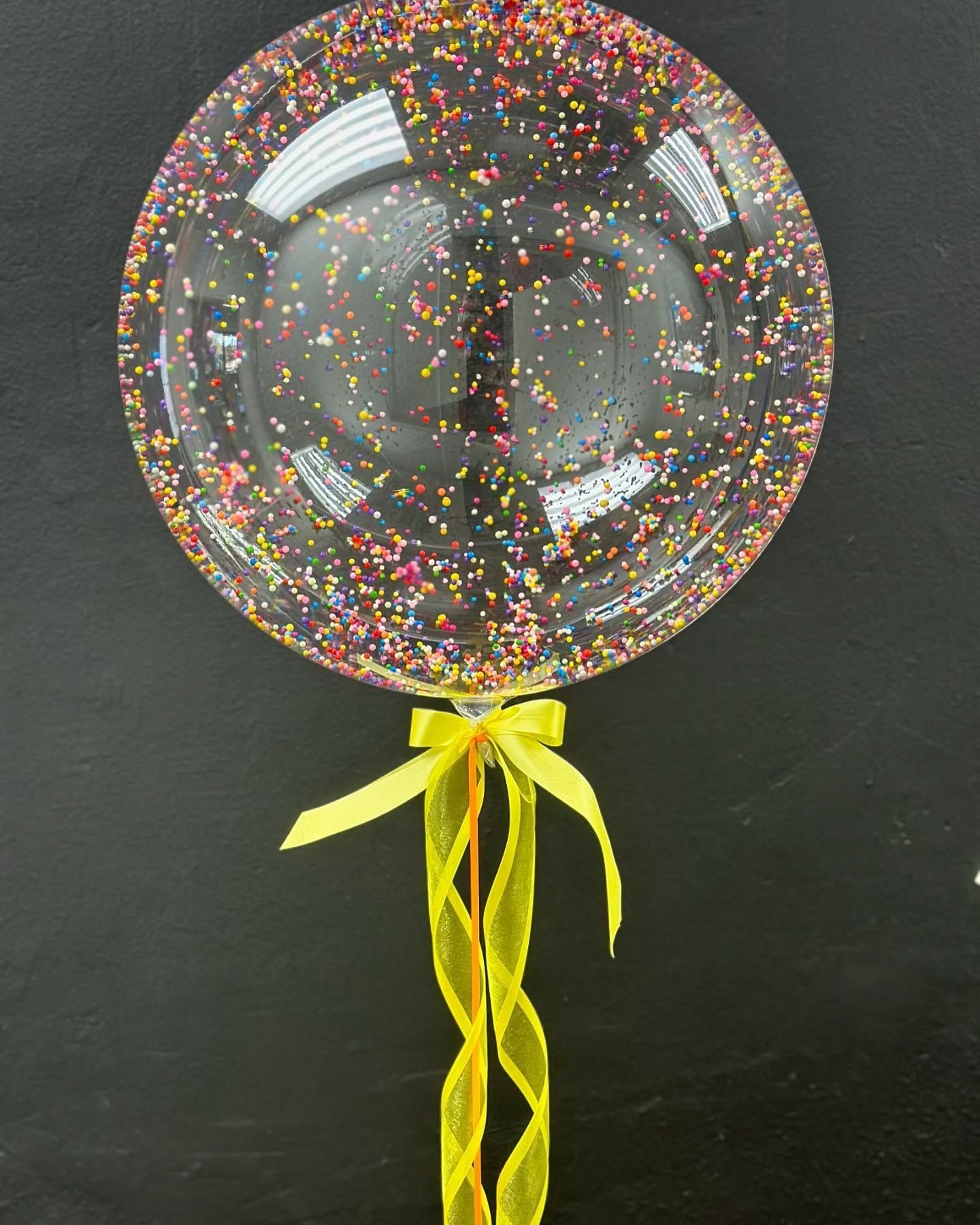 A clear balloon filled with confetti and a yellow ribbon on a stick.