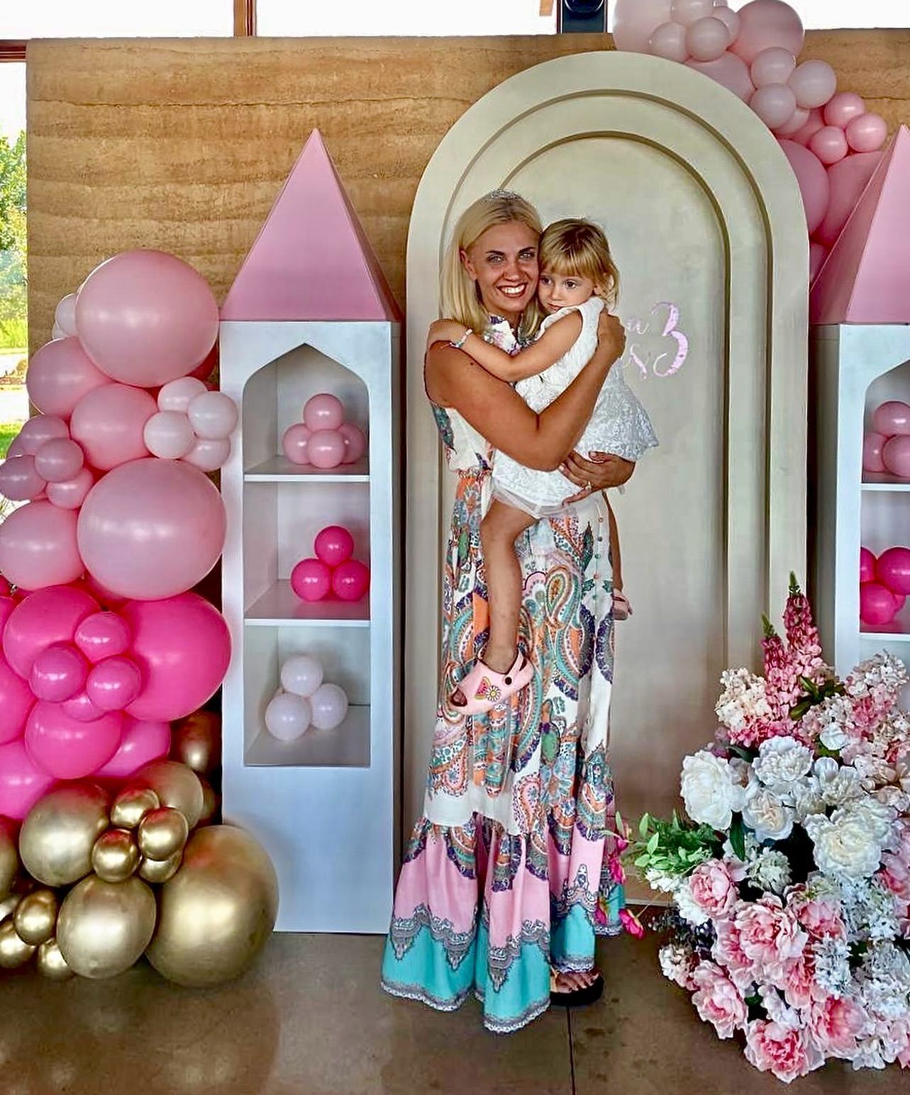 A woman is holding a little girl in front of a wall decorated with pink balloons.