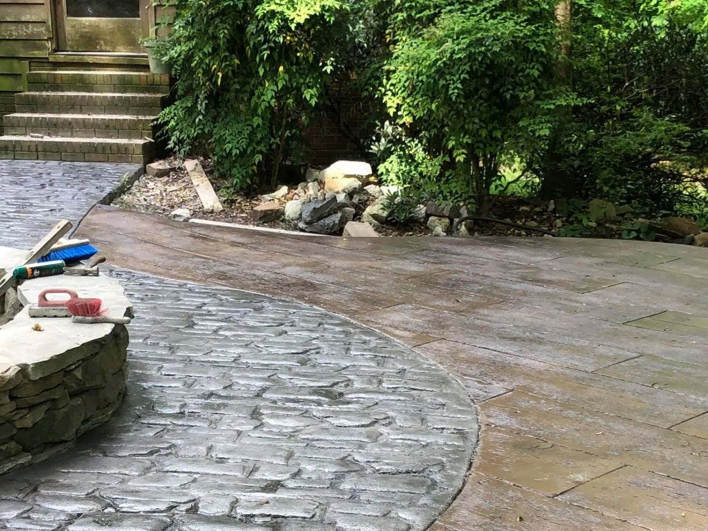 Stone patio with a curved border, leading to a path and garden with lush greenery and stairs.