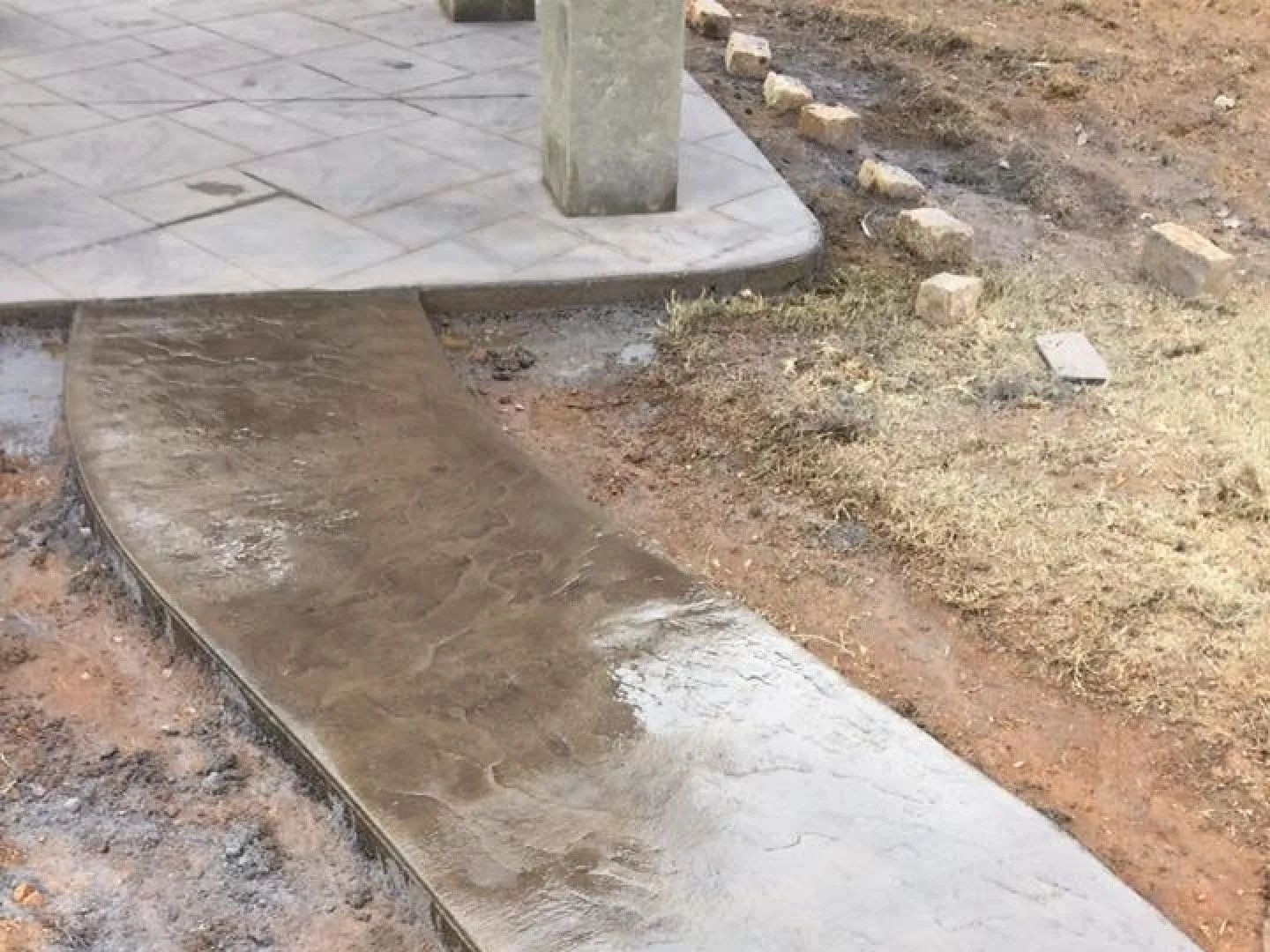 Concrete walkway leading from a patio, under a concrete column, through dirt and grass.