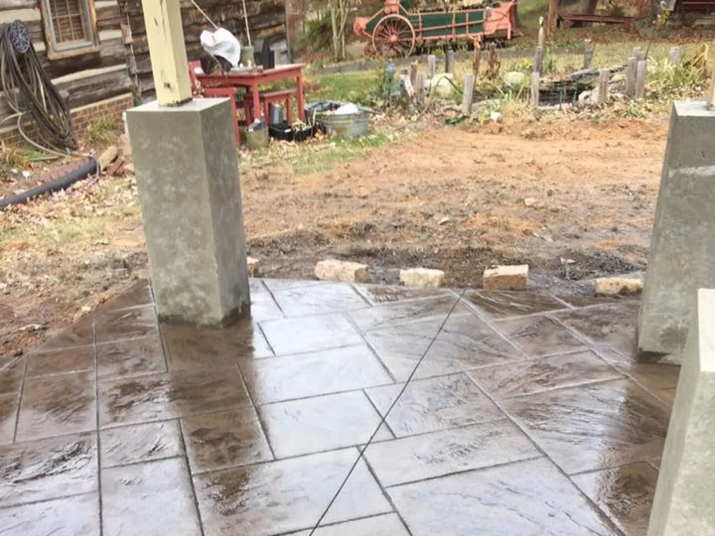Concrete patio under construction with square and angled patterns. Two pillars are visible. Outdoor setting.