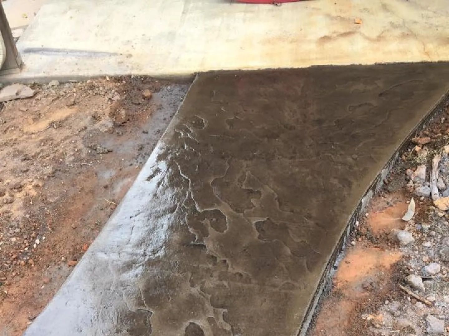 Concrete walkway, dark brown, textured surface, next to dirt and a light-colored building.