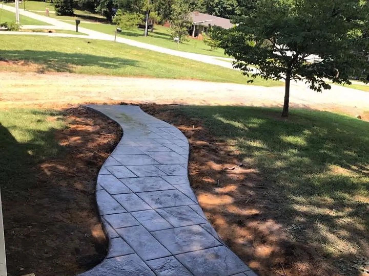 A winding stamped concrete walkway in a yard, bordered by brown soil and green grass.
