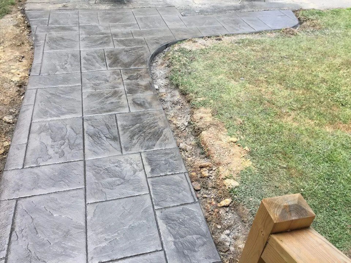 Gray stamped concrete walkway curving along a grassy area, with exposed dirt along the edge.