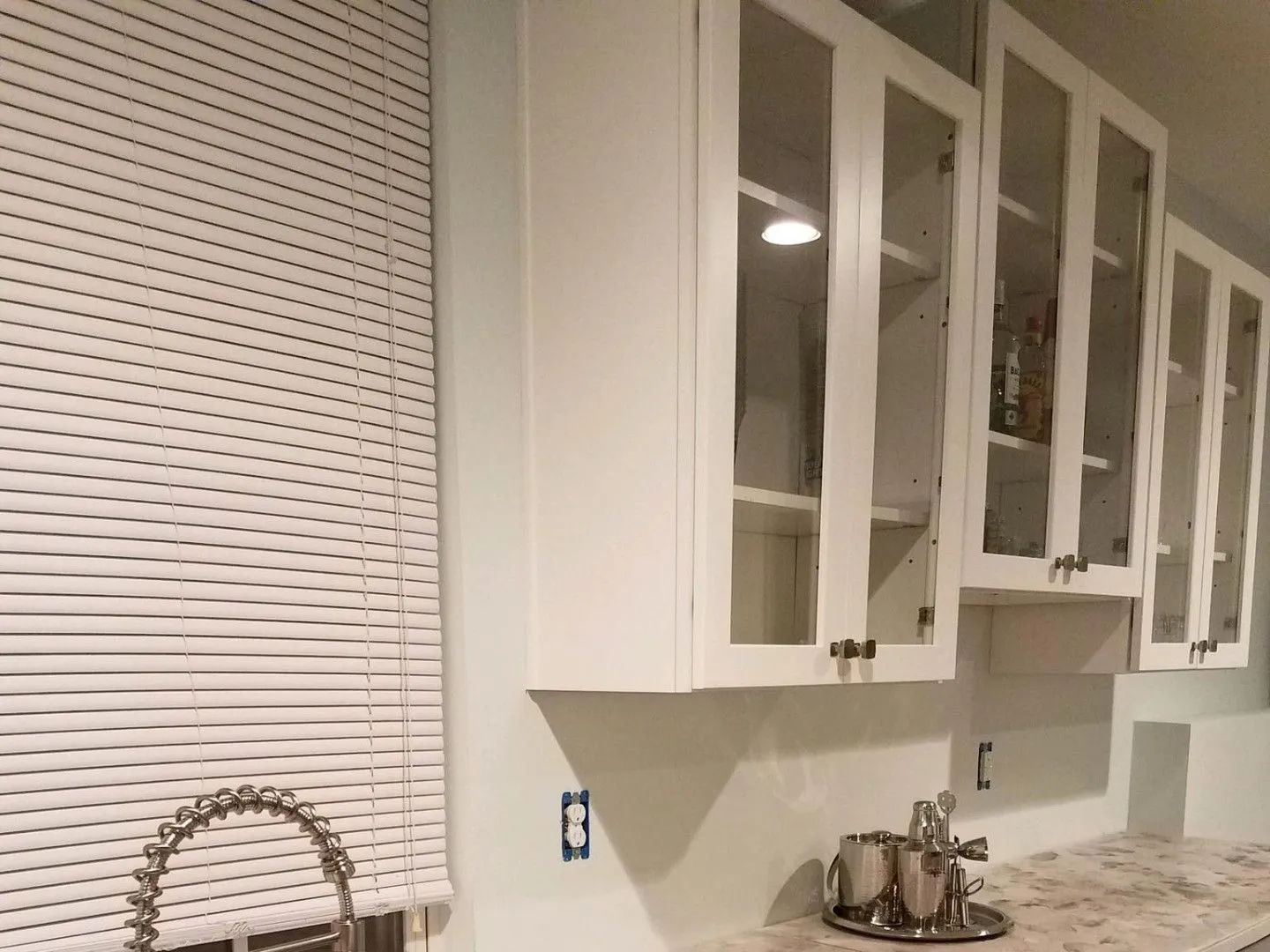 White kitchen cabinets with glass doors over a countertop, blinds on the window, and a silver faucet.