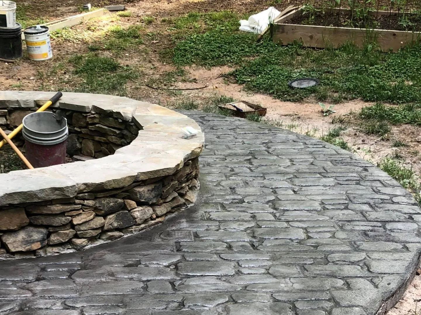 Fire pit with stone surround next to a concrete patio with brick pattern, outdoors.