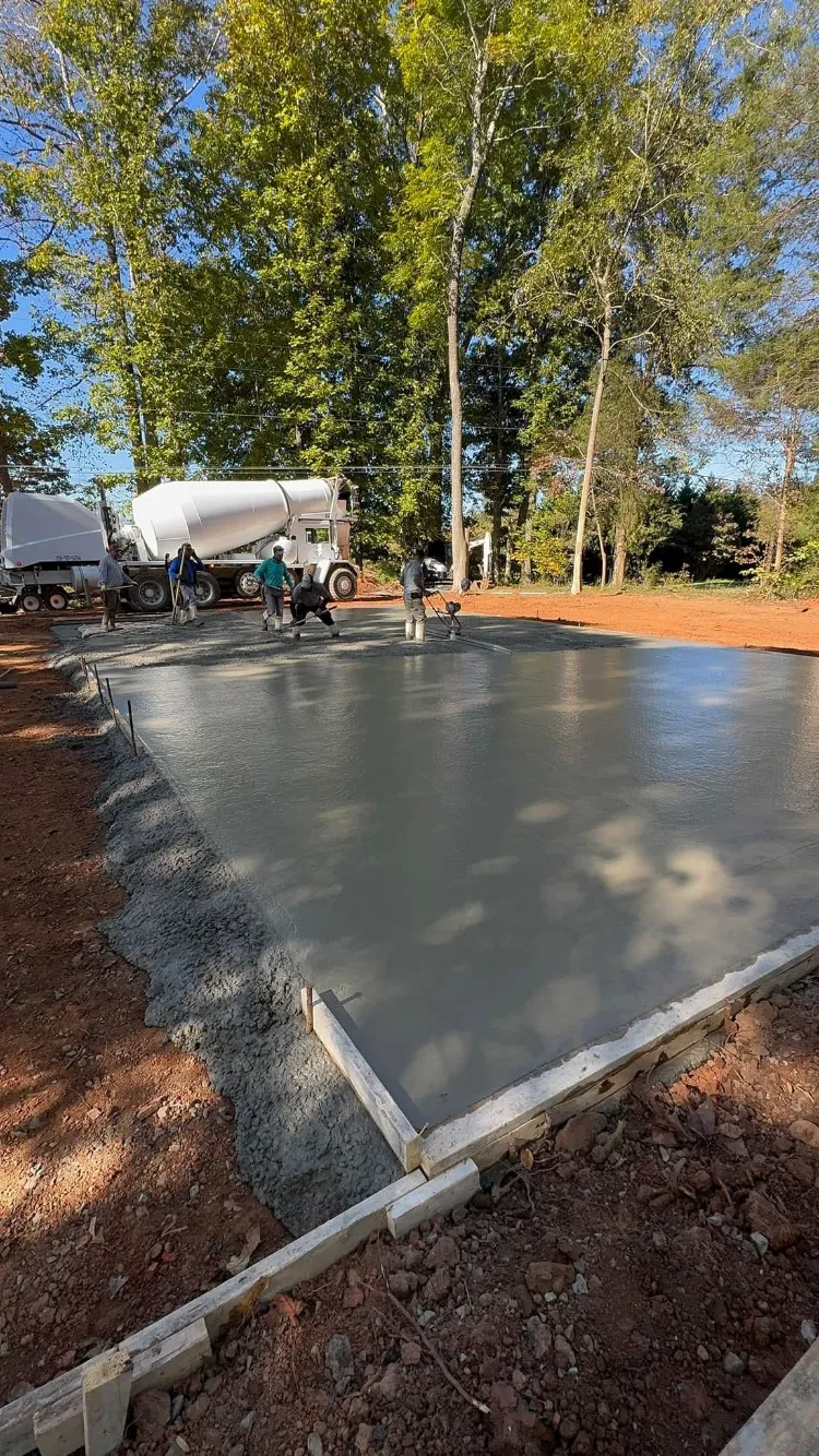 Workers pouring and smoothing fresh concrete for a rectangular foundation outdoors, with a cement truck in the background.